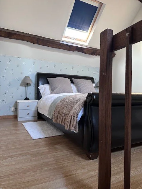 Bedroom with a pitched ceiling, skylight, bed with beige and white bedding, black headboard, nightstand, lamp, wooden floors, and a blanket draped over the bed.