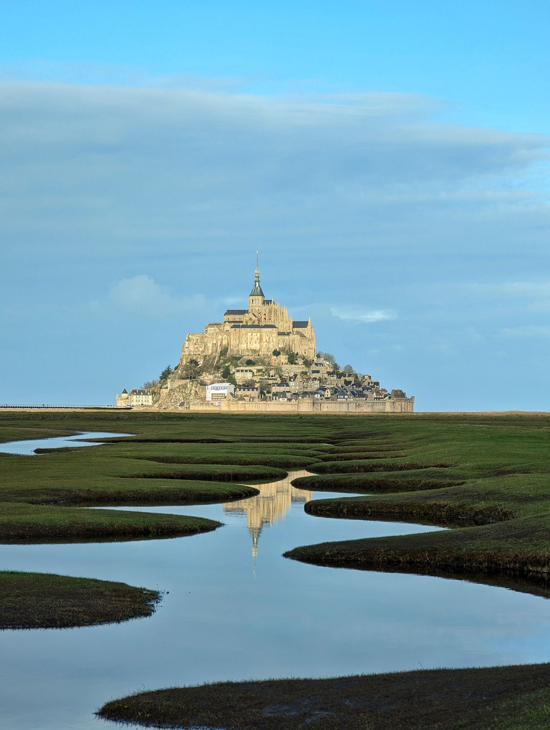 View of Mont Saint-Michel, a historic island monastery in Normandy, France, with a reflecting waterway in the foreground under a partly cloudy blue sky.