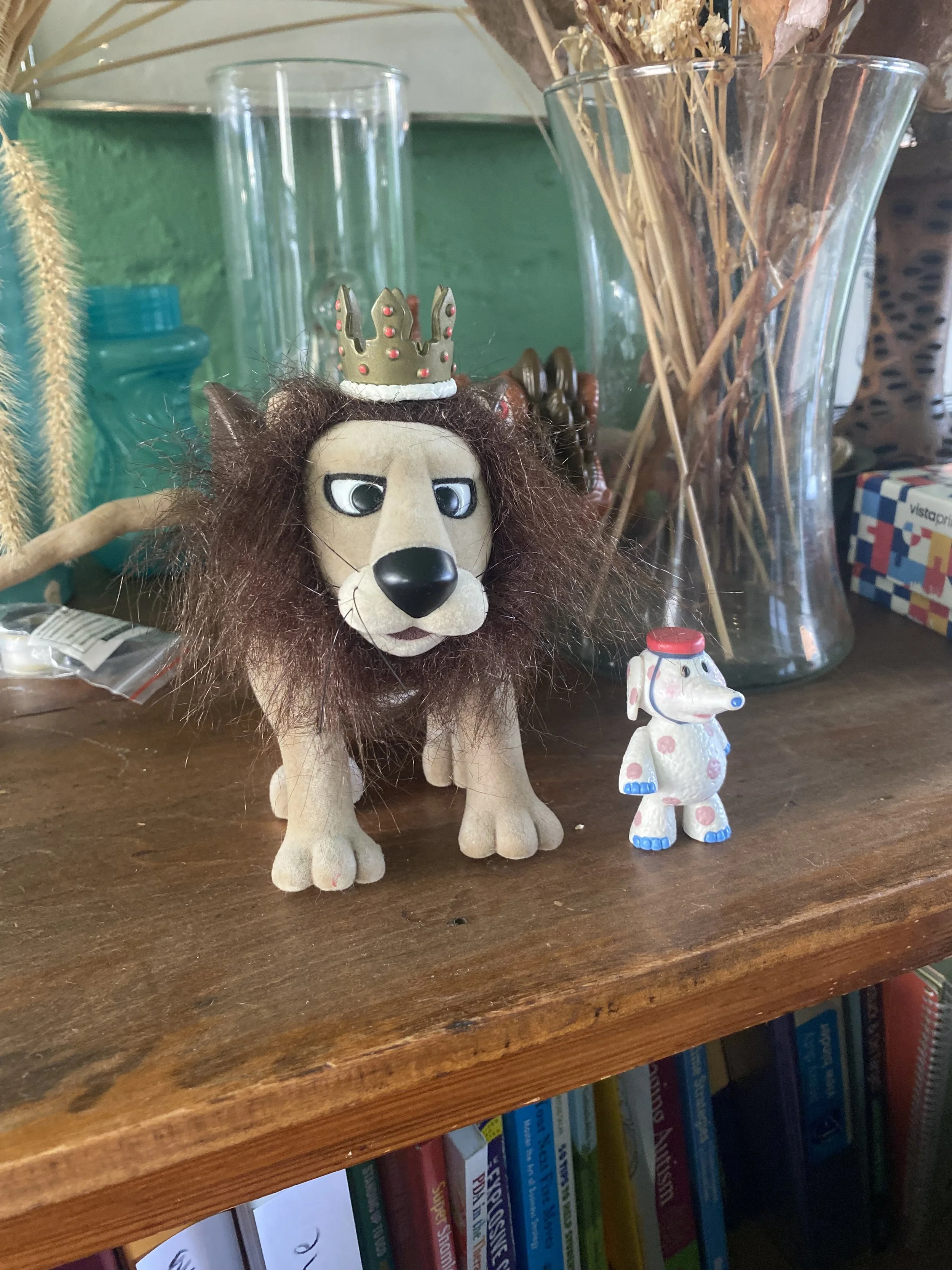 A collection of small toys on a wooden shelf, including a plush lion with a crown on its head and a small plastic dog figurine with painted spots, surrounded by glass vases and dried plants.