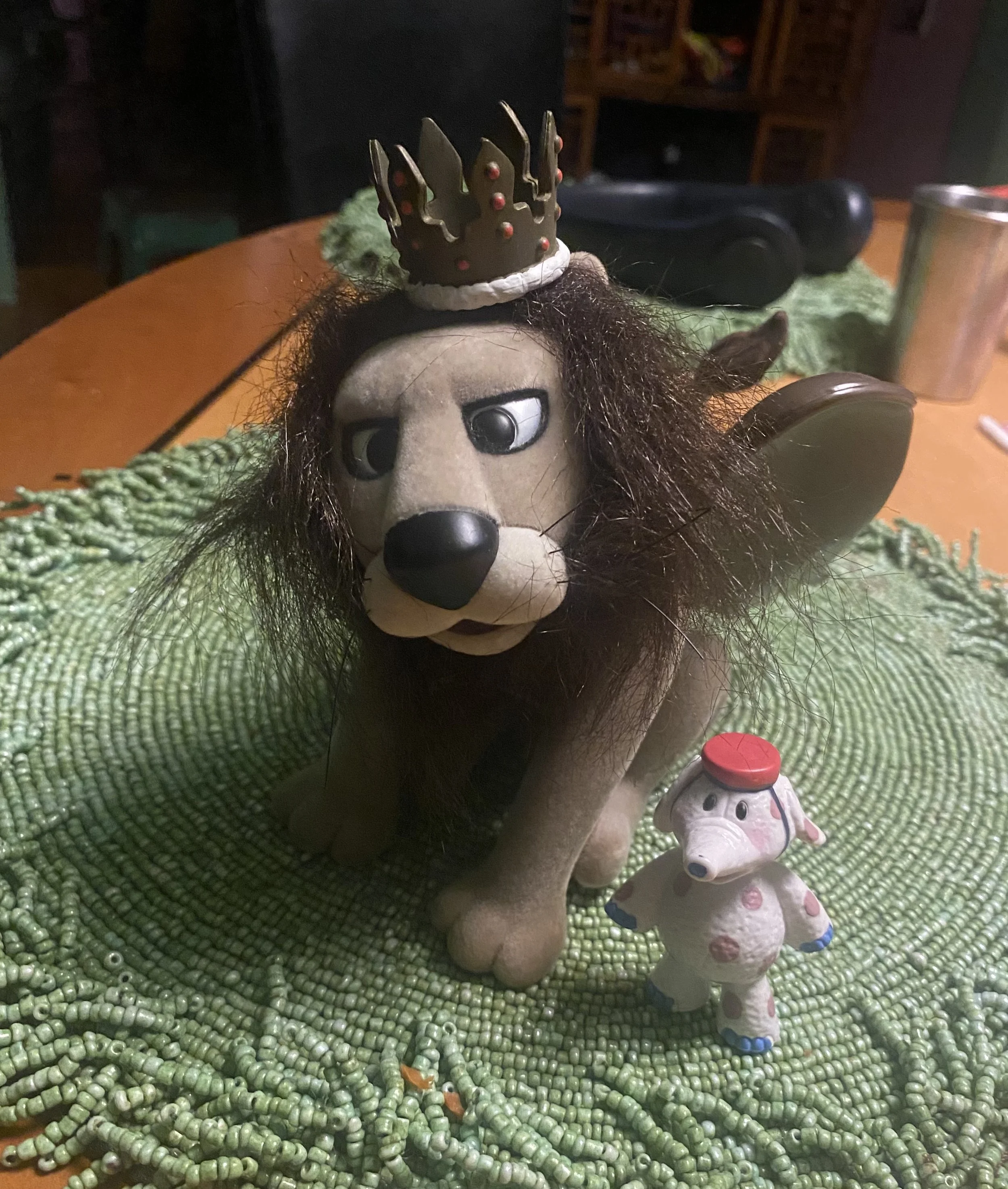 A plush toy of a dog with large eyes and long dark hair, wearing a small black crown decorated with red beads, sitting on a green beaded surface next to a small figurine of a cow with pink spots and a red and white hat.