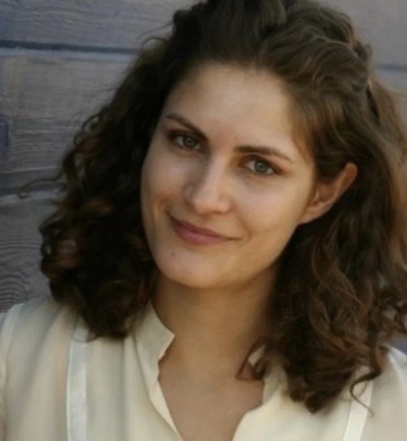 Close-up portrait of a woman with wavy brown hair, blue eyes, and a gentle smile, wearing a light-colored blouse, sitting outdoors near a wooden wall.