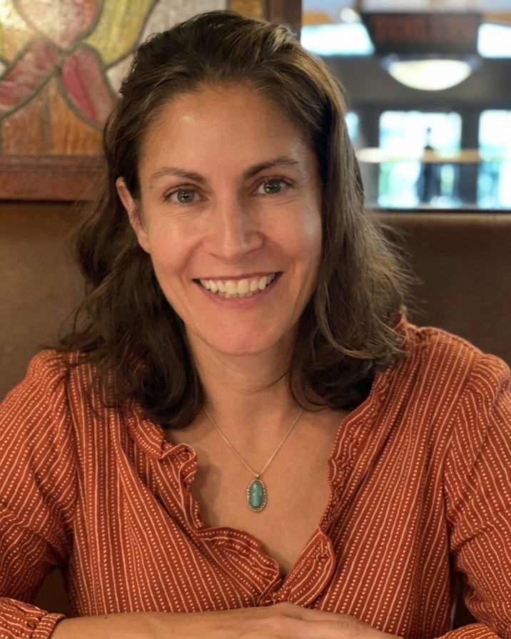 Smiling woman with brown wavy hair wearing an orange and white striped blouse and a turquoise pendant necklace, sitting in a restaurant or café.