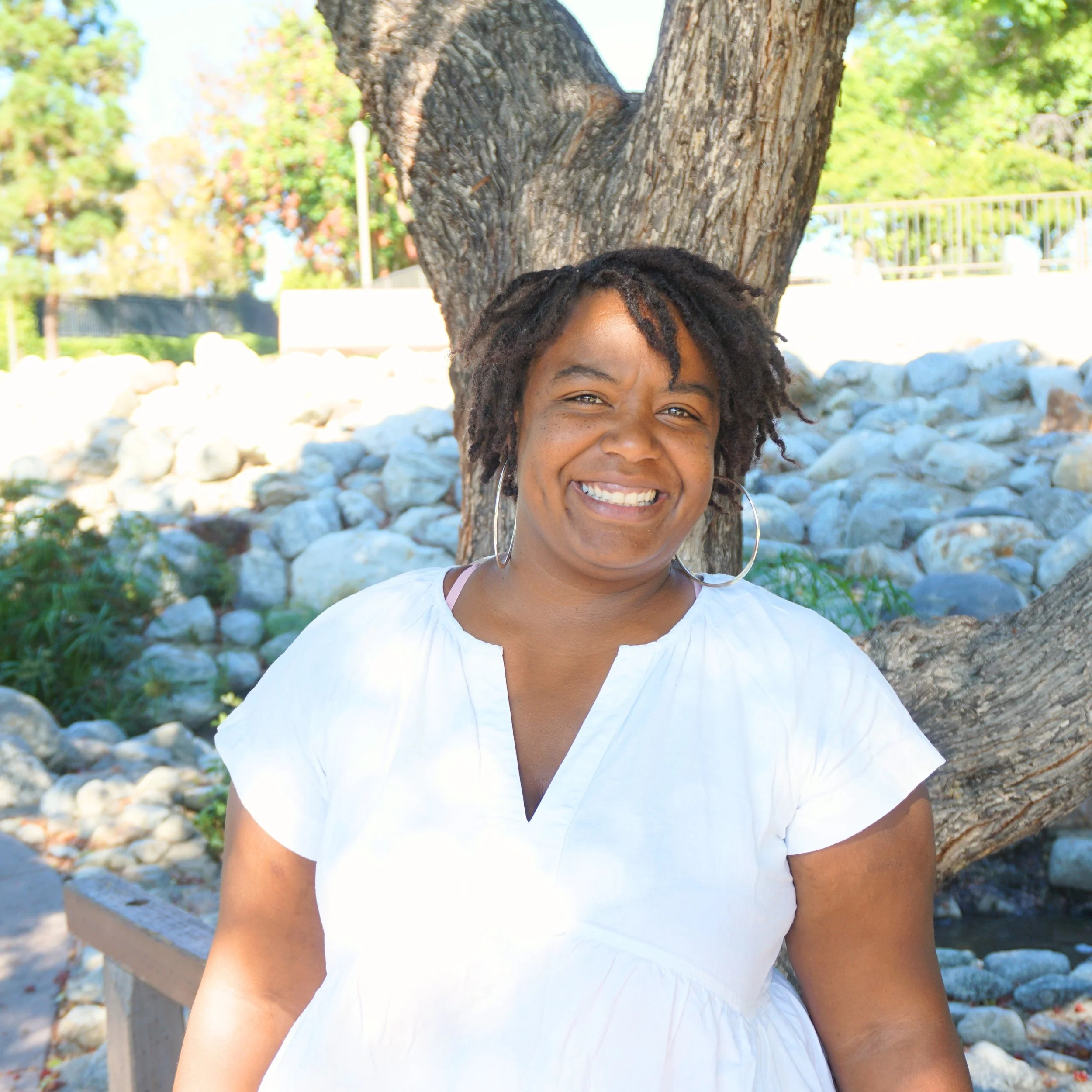 The image shows a woman smiling, standing outdoors in front of a large tree with textured bark. The background includes a stone-lined area, rocks, trees, and a metal fence, suggesting a park or garden setting on a sunny day.