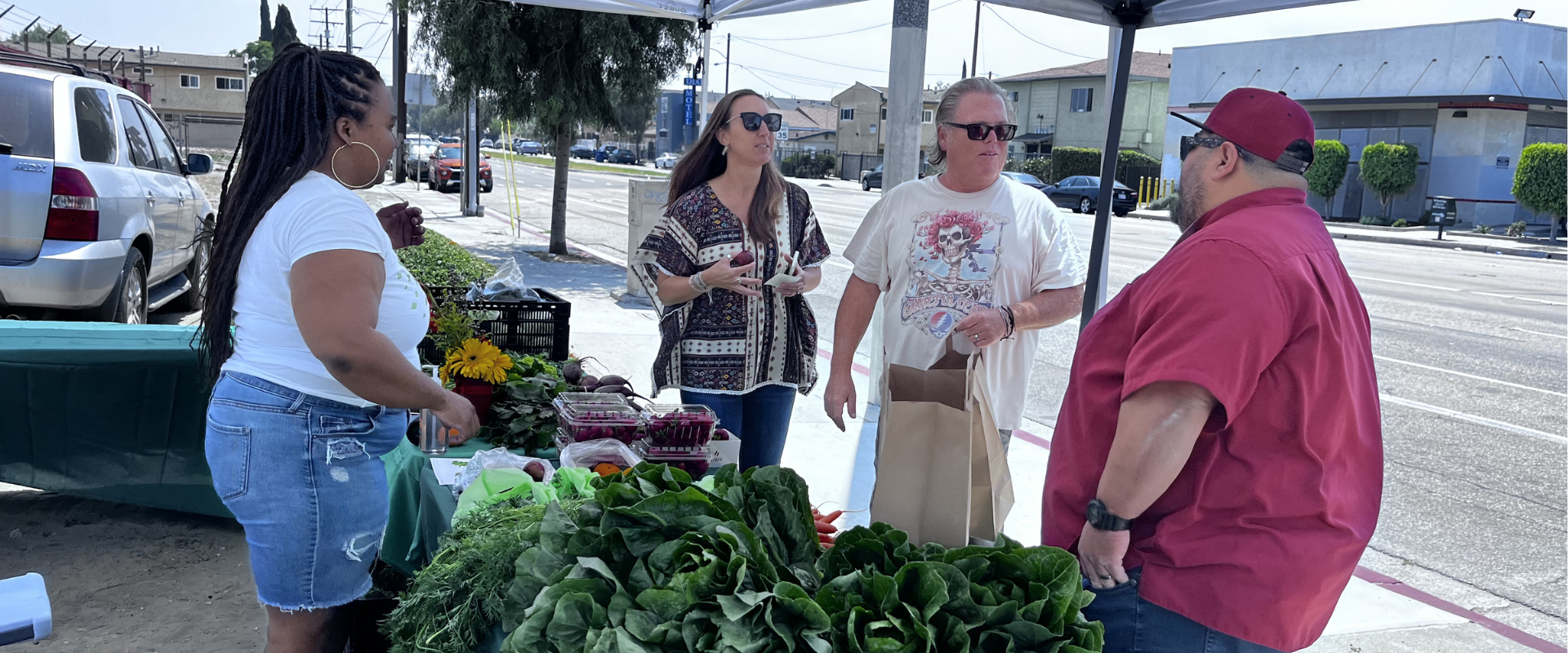 Four people talking at an outdoor farmer's market stand with fresh vegetables and flowers, along a city street with parked cars and buildings in the background.