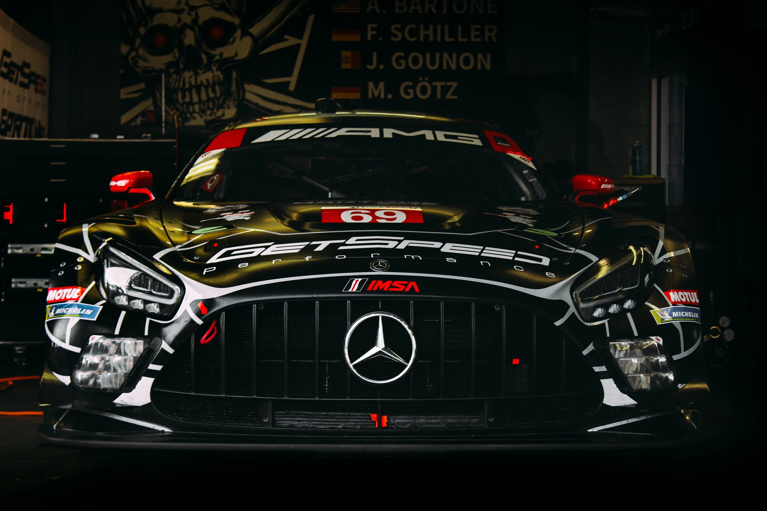 A black racing car with Mercedes-Benz logo, decorated with sponsor logos including Petronas, Michelin, and Motul, parked in a garage or workshop. The background features a wall with a large skull artwork and a list of names and flags.