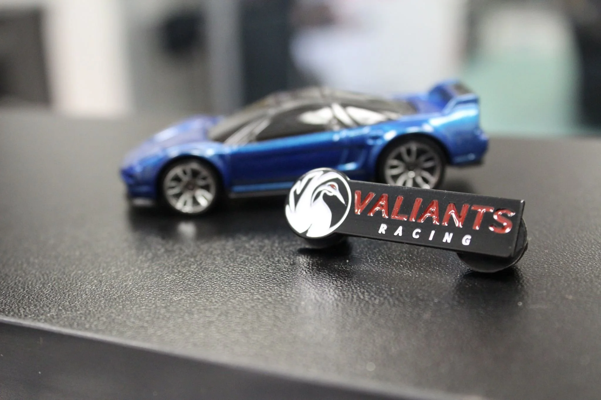 A small blue toy sports car placed on a black surface with a 'Valliants Racing' badge in front of it. The background is blurred.
