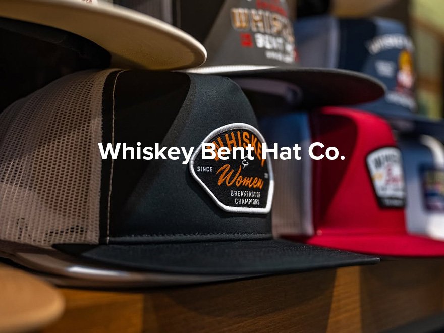 A display of several trucker hats with various designs. One hat in the foreground has a patch that reads 'Whiskey Bent Hat Co.' and 'Women Breakfast Champions.'