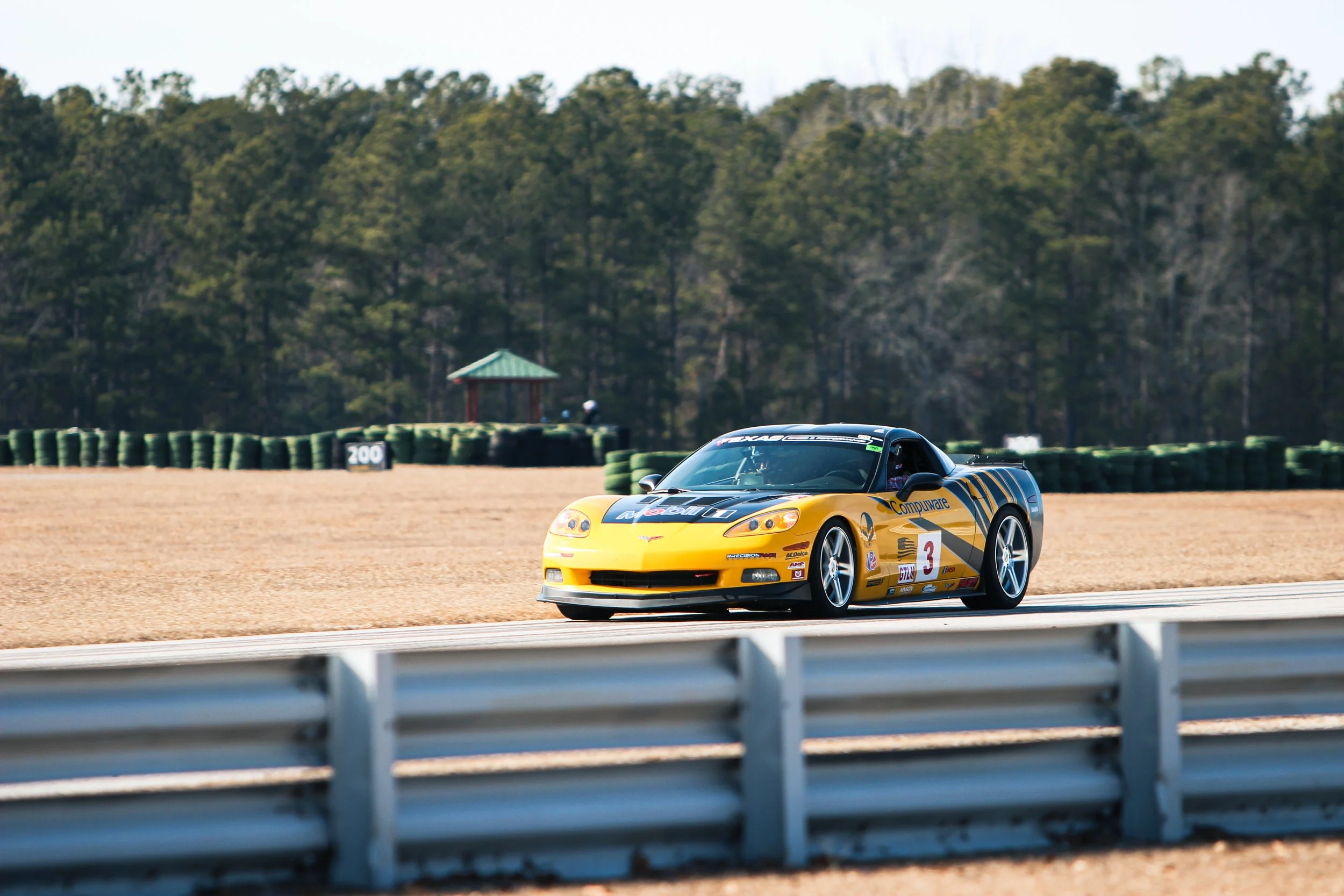 A yellow racing car with sponsorship decals driving on a racetrack with green tire barriers and a wooded background.