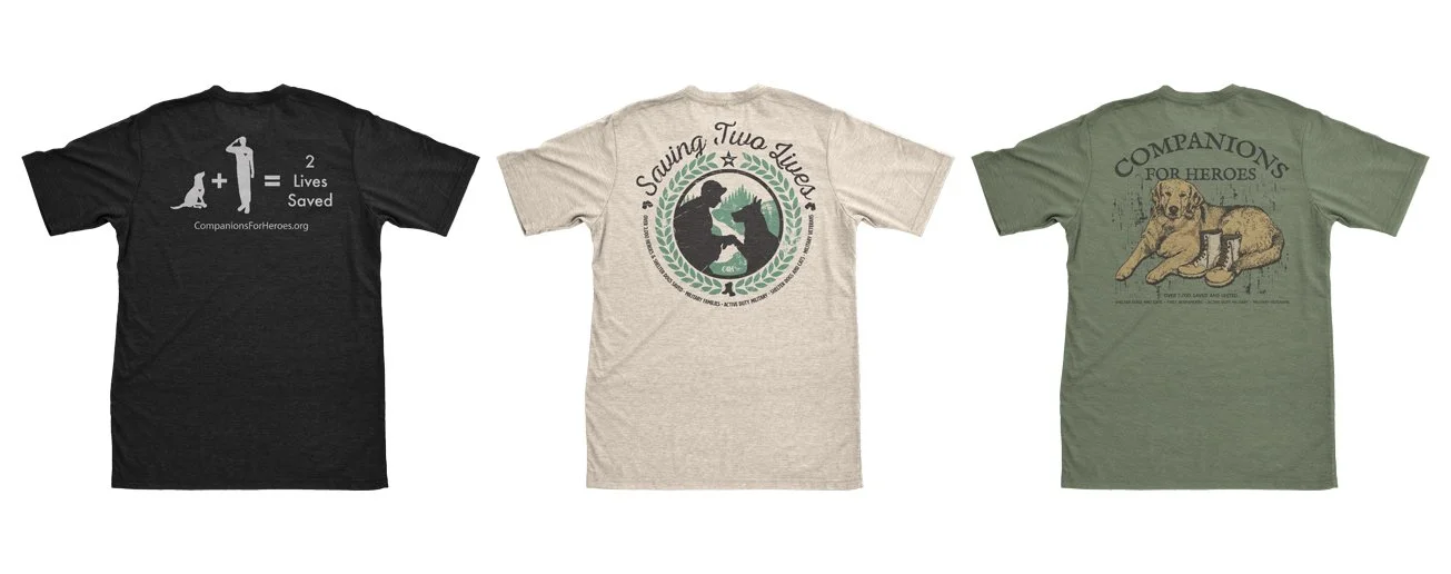 Three T-shirts displayed side by side, each with a different animal rescue message and graphic. The first shirt is black with a graphic of a dog and human silhouette, stating "2 Lives Saved." The second shirt is light gray with a design of two dogs in a circle, reading "Saving Two Lives." The third shirt is green with a graphic of a dog lying down and the text "Companions for Heroes."