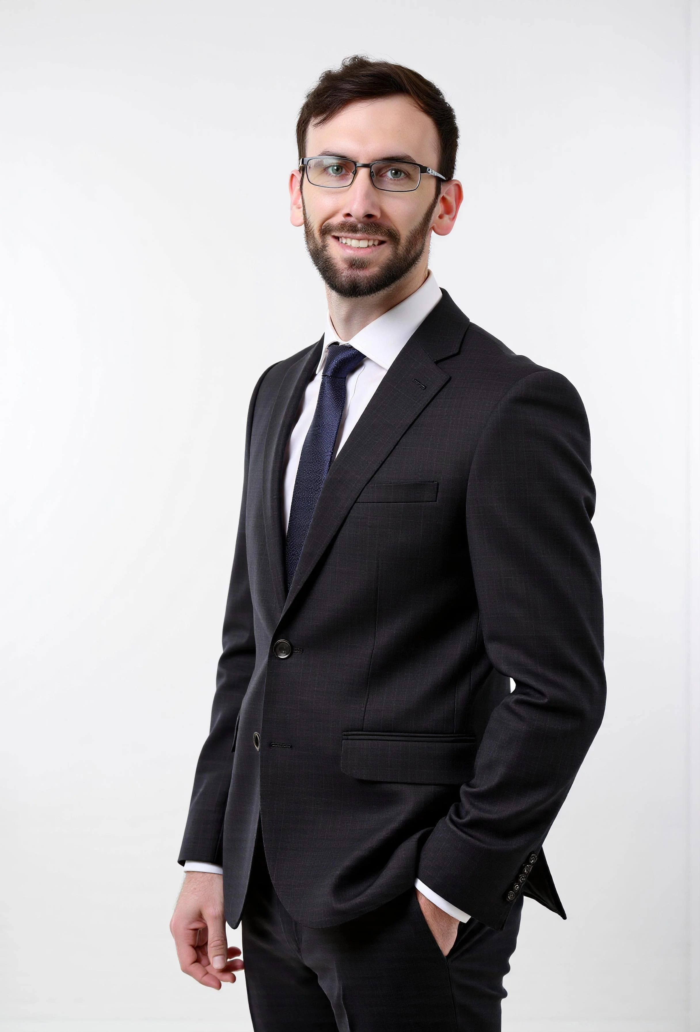 A man wearing glasses and a dark suit with a white shirt and dark tie, standing against a plain white background, smiling and looking at the camera.