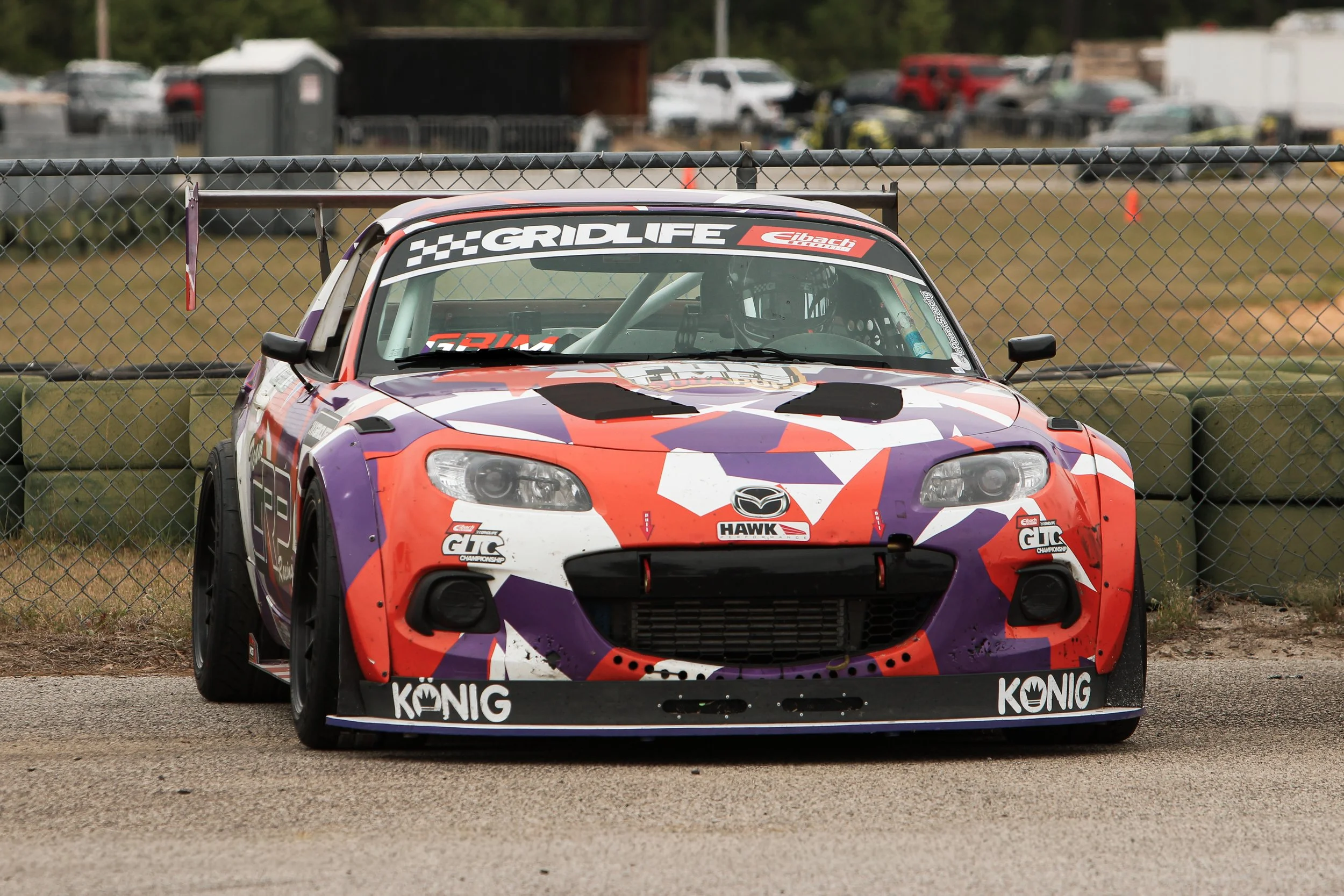 A racing car with a colorful purple, orange, and white geometric design on the front, behind a chain-link fence at a race track.