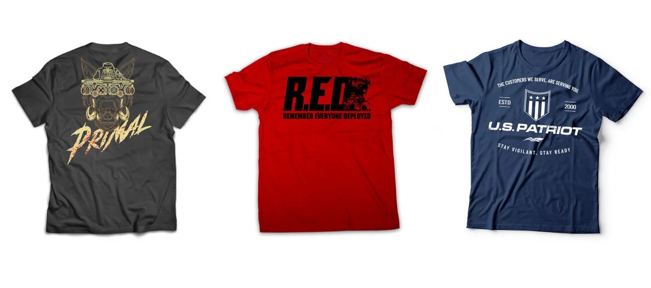 Three T-shirts laid flat, each with different printed designs and colors. The first is a dark gray shirt with a gold graphic of a armored tank and horned beast, and word "PRIMAL" in cursive. The second is a red shirt with bold black text "R.E.D" and the phrase "REMEMBER EVERYONE DEPLOYED" underneath, along with a black graphic of a soldier. The third is a blue shirt with white text and graphics, including a shield, stars, and the phrases "U.S. PATRIOT," "STAY VIGILANT, STAY READY," and "THE CUSTOMERS WE SERVE, ARE SERVING YOU!"