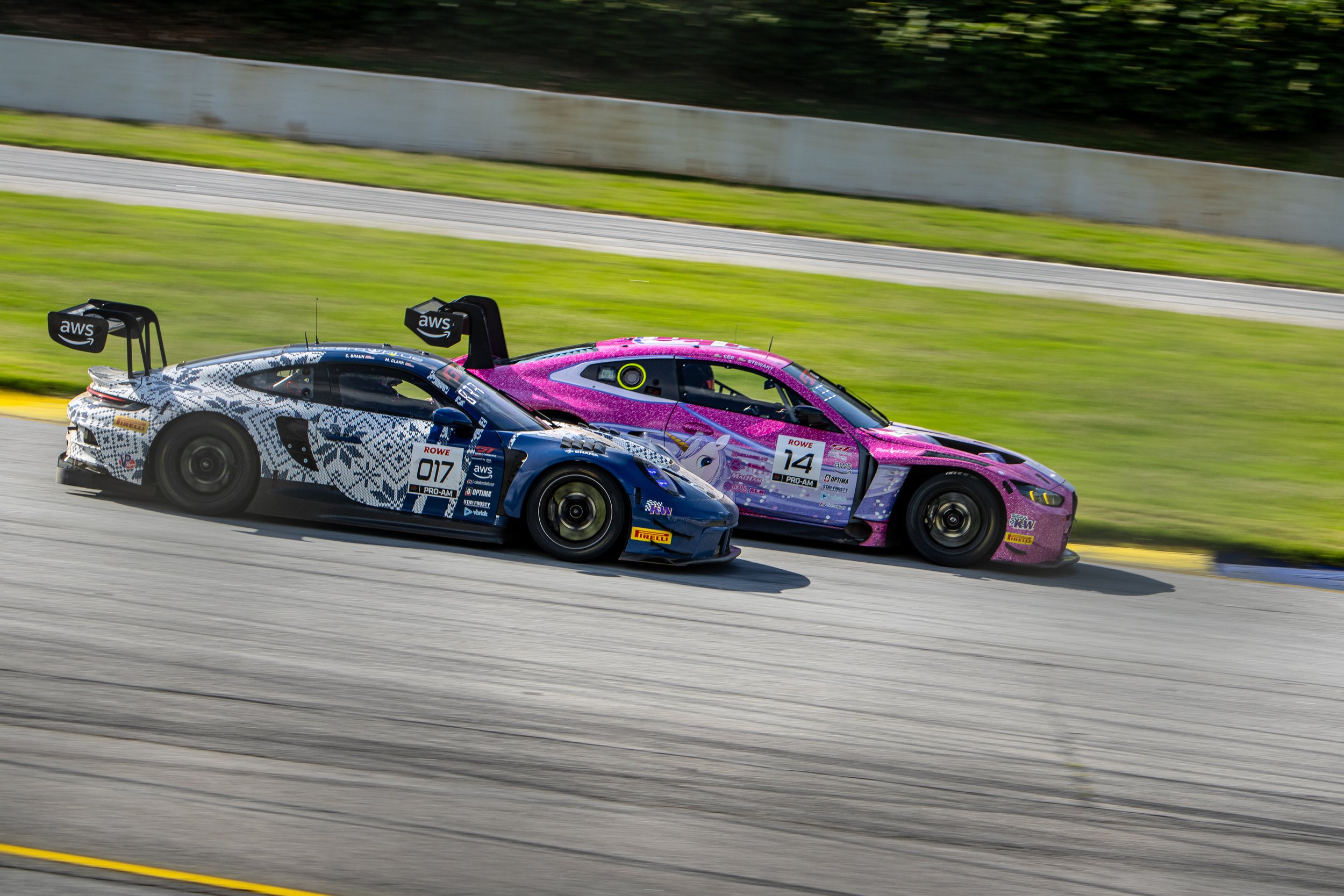 Two race cars, one with a black and white patterned design and the other with pink and purple graphics, are speeding along a race track with blurred motion in the background.