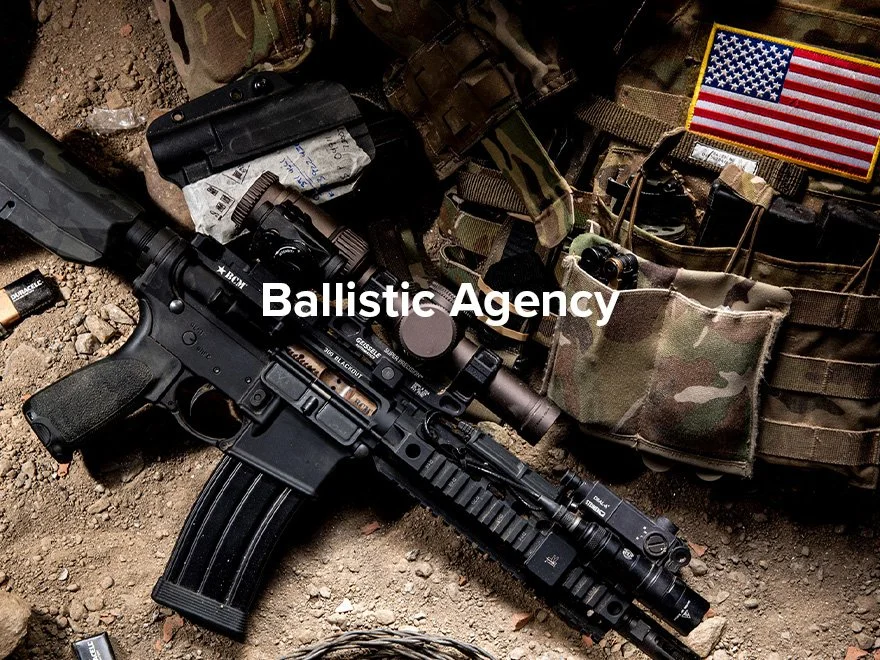 A black rifle with mounted scope and tactical accessories lying on dirt ground, surrounded by camouflage military gear, a patch of the American flag, and other tactical equipment.