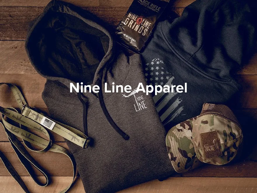 Clothing and accessories, including a gray hoodie with 'Nine Line' logo, a black hoodie, camouflage-patterned pouch, and a dog leash with 'Nine Line' branding, all displayed on a wooden surface.