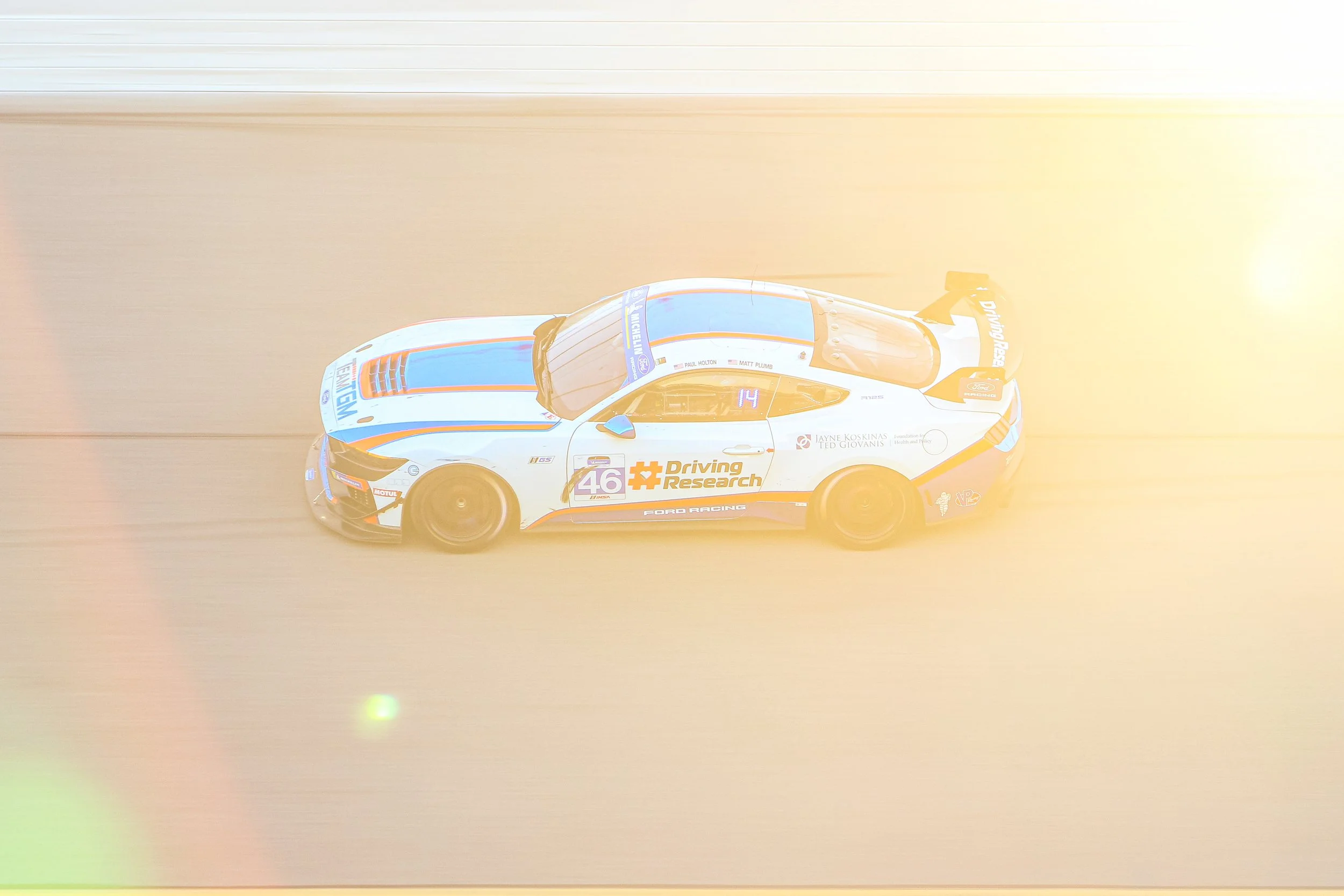 A photograph of a race car on a track, taken from above, with sunlight glare on the top right corner of the image.