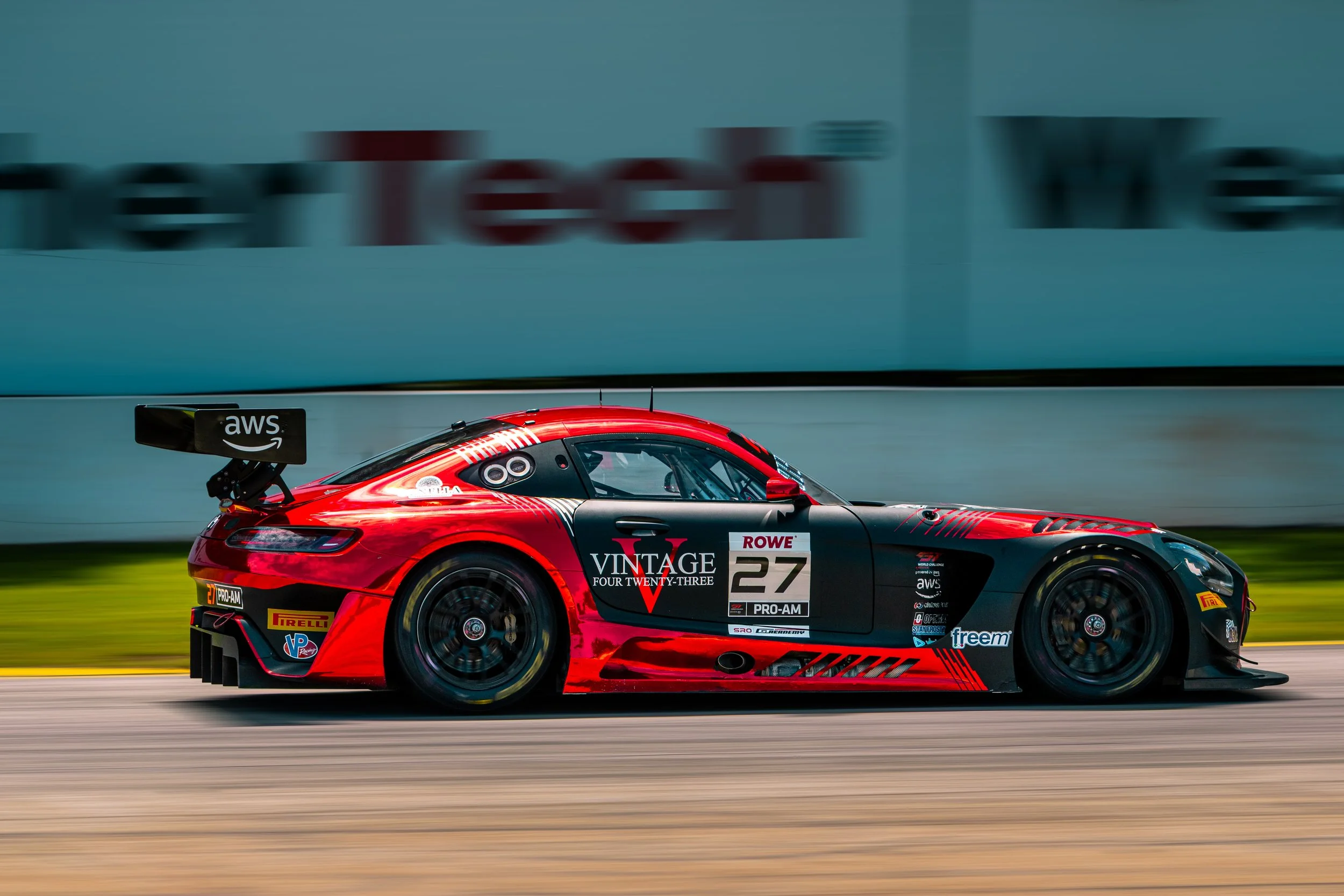 A red race car with various sponsor logos, including AWS and Pirelli, moving at high speed on a race track.