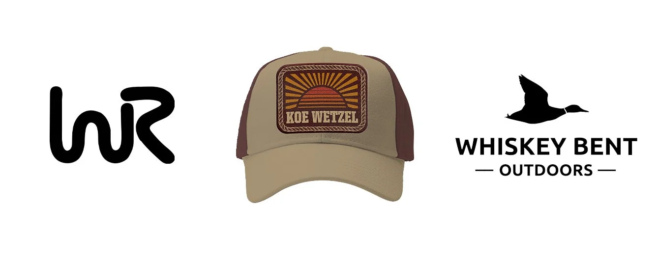 A beige and brown trucker hat with a patch that reads 'Koe Wetzel' and an illustration of a sunrise. To the left is a black stylized letter 'W', and to the right is a logo with a flying bird silhouette, the text 'Whiskey Bent Outdoors', and decorative lines.