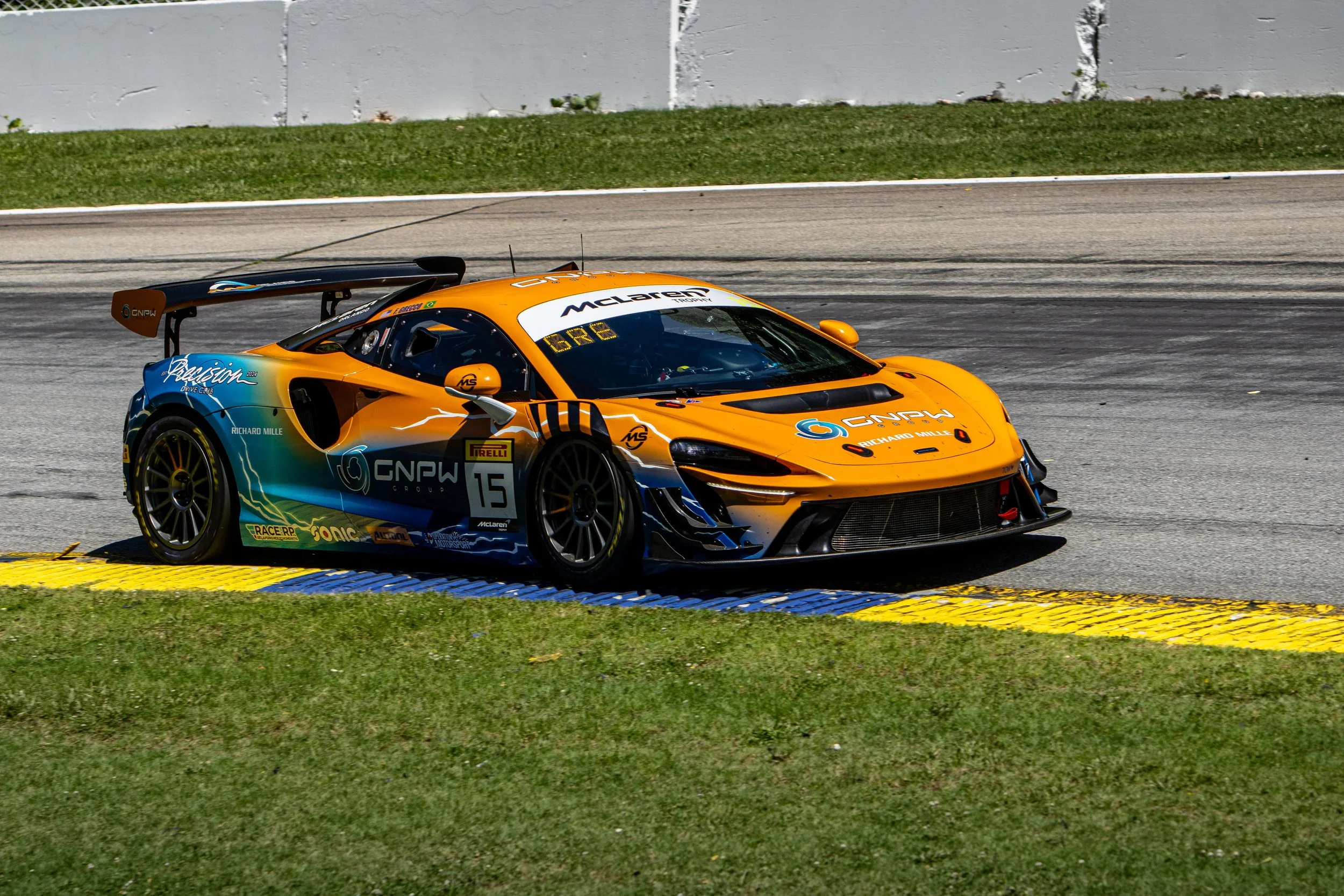 A bright orange racing car on a race track, with various sponsor logos and the number 15 on the side, positioned near a curb with yellow and blue markings, with a gray wall and green grass in the background.