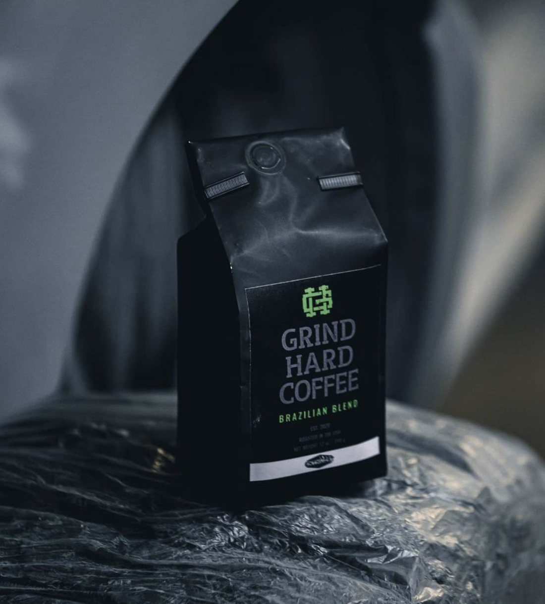 A black bag of Grind Hard Coffee Brazilian Blend placed on shiny plastic-wrapped surface with a metallic background.