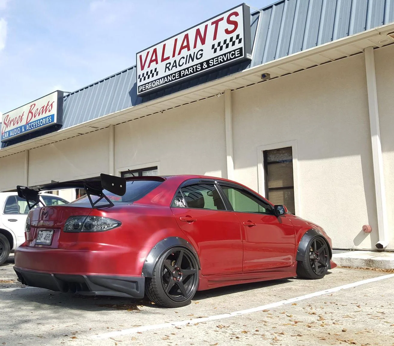 A red modified car with black rims and a large rear spoiler parked outside an auto parts store named Valiant Racing with signs for car audio and accessories and performance parts and service.
