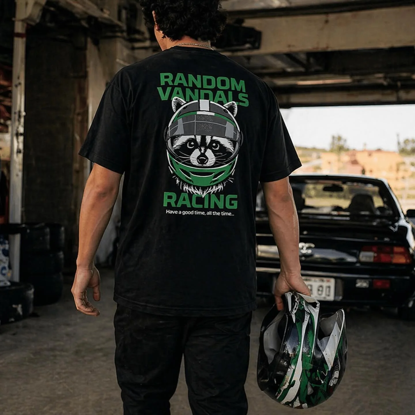 Person in black T-shirt holding racing helmet, standing in garage, facing away, with black sports car in background