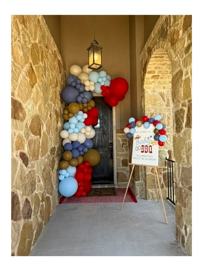 Decorative balloon arrangement in front of a black front door, with a welcome sign for a baby shower celebration for Kaley and Andrew, featuring red, blue, white, gray, and gold balloons.