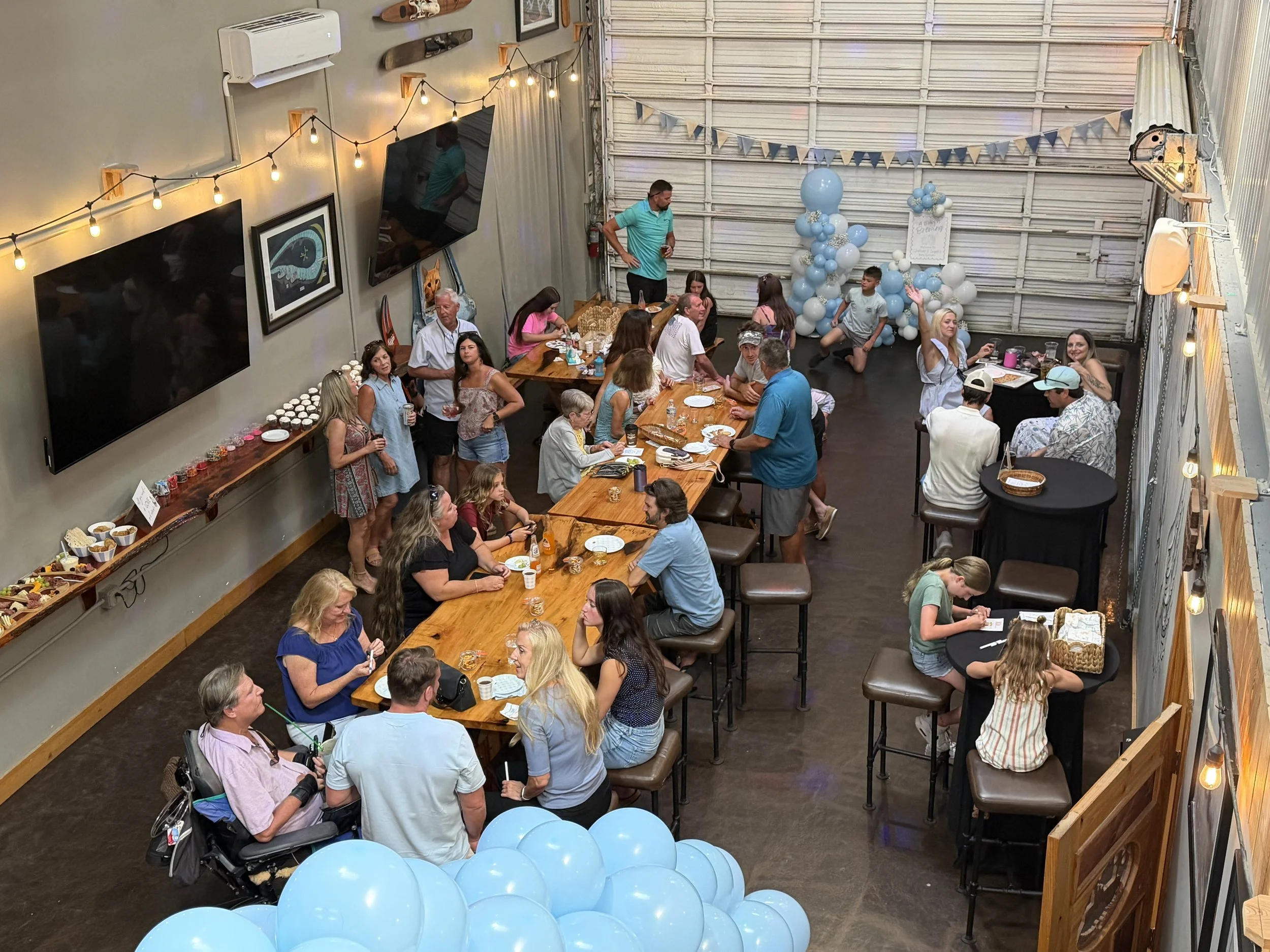 Indoor party scene with many people sitting and standing at long wooden tables, blue balloon decorations, and string lights hanging on the walls.