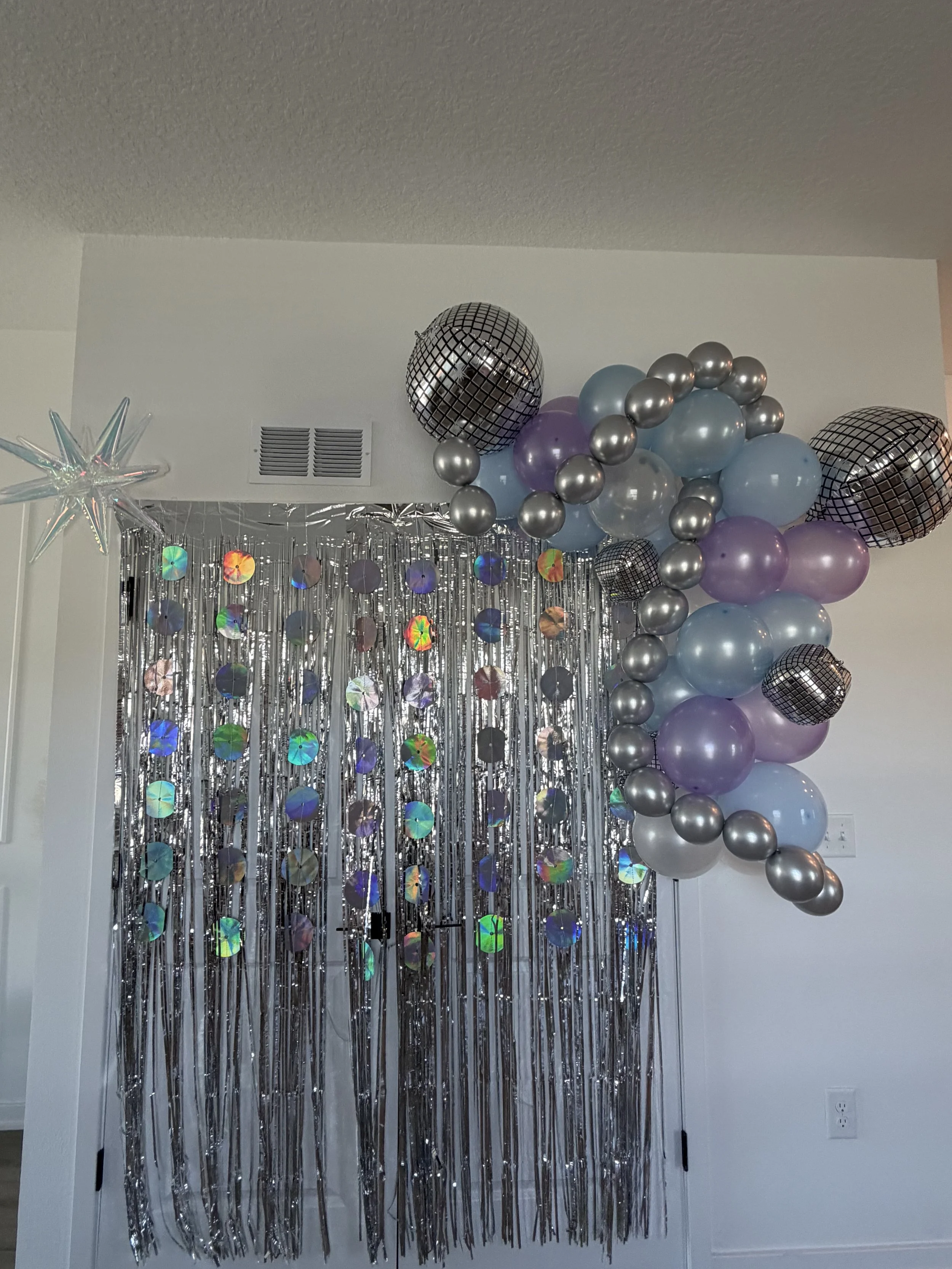 Decorative party backdrop with silver tinsel curtains, holographic discs, and a balloon arrangement featuring silver, purple, and blue balloons with some metallic black balloons and a star-shaped foil balloon.