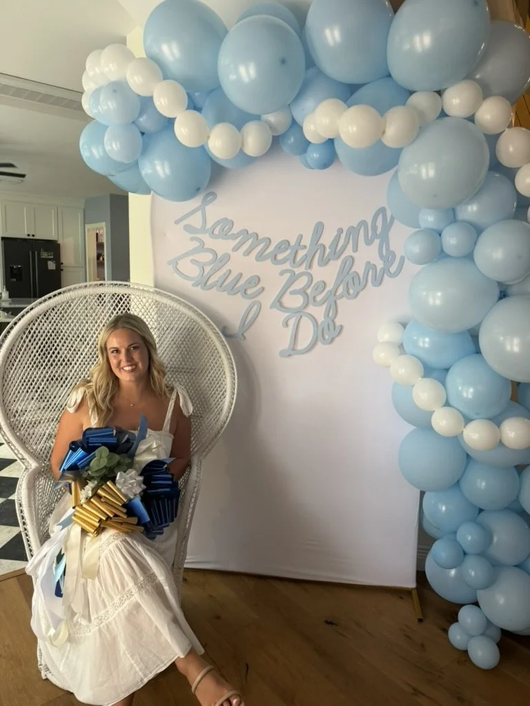 Bridal shower backdrop featuring a white vintage peacock chair, custom balloon garland, and gold arch backdrop by Pink Palm Events Co.