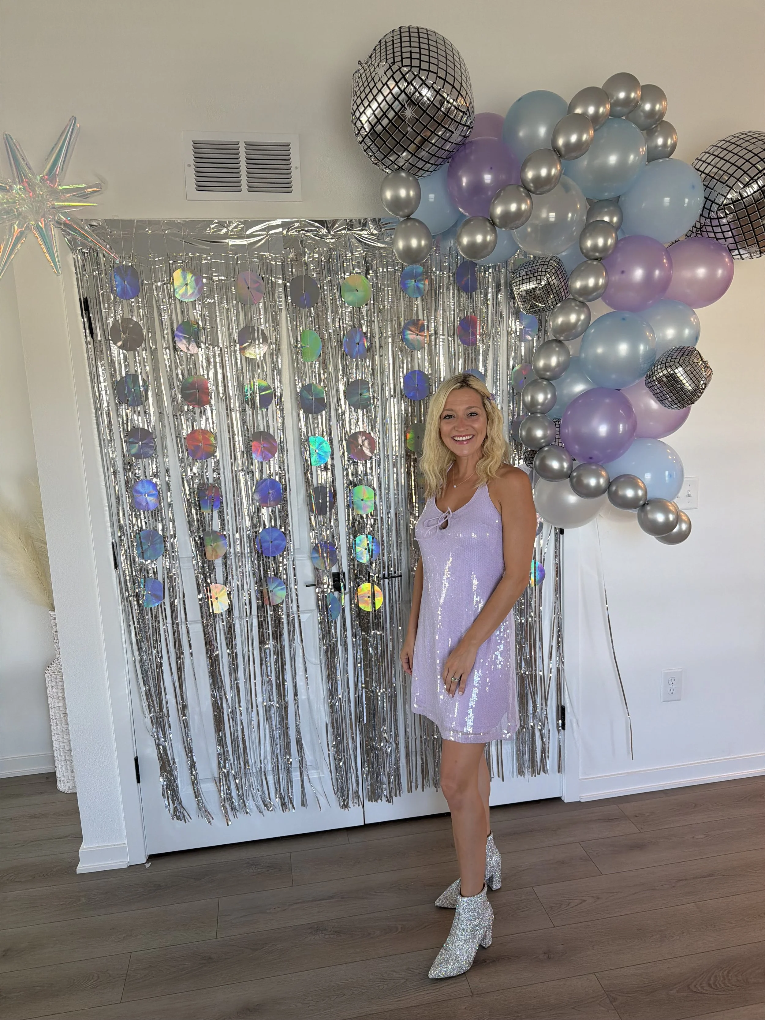 A woman in a sequined purple dress and glittery platform boots standing in front of a silver and pastel balloon and foil decoration.
