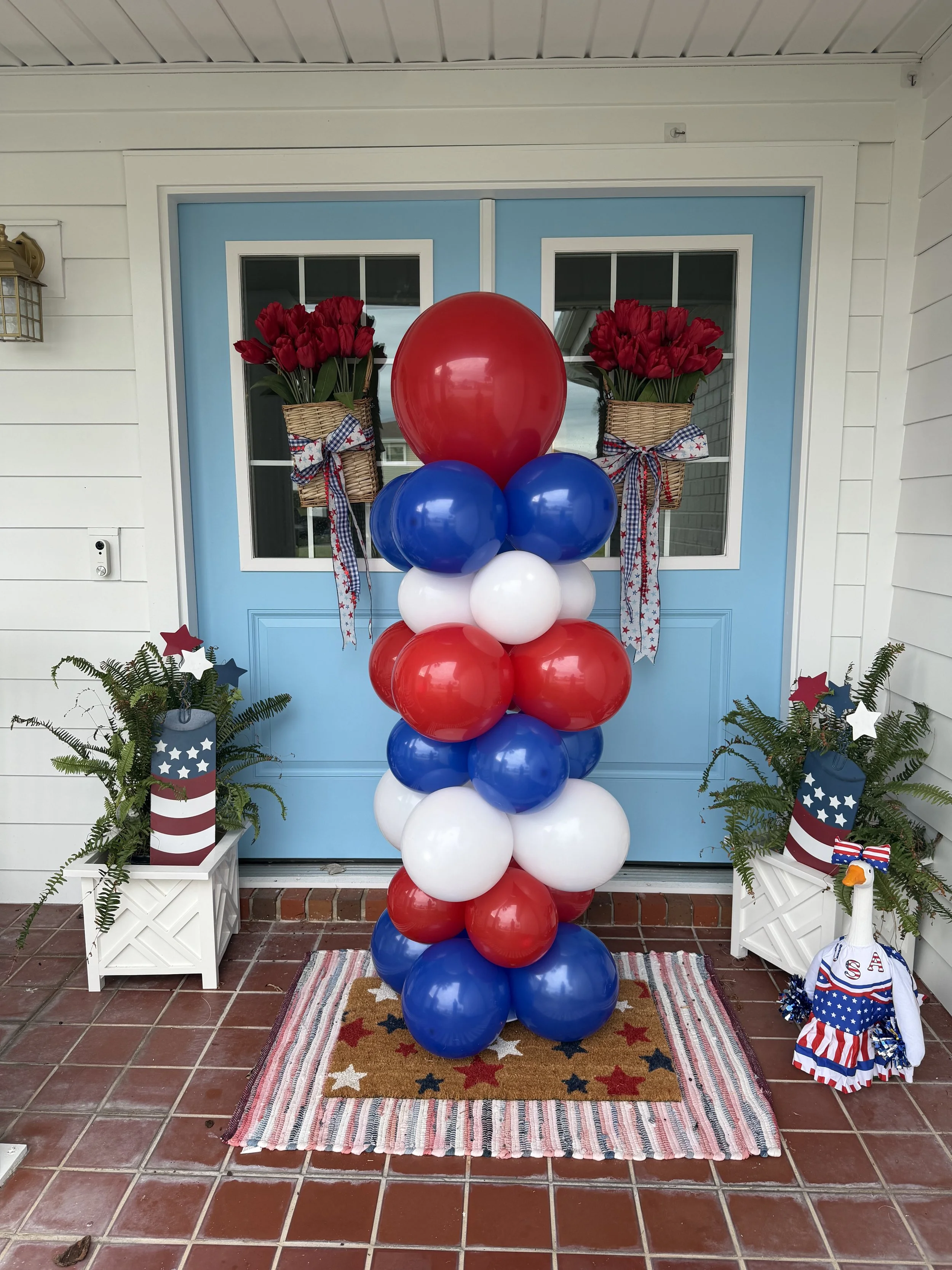 Patriotic outdoor door display with red, white, and blue balloons arranged vertically in front of a blue door with two small windows. On each side, there are white planters with ferns decorated with red, white, and blue star-patterned patriotic decor