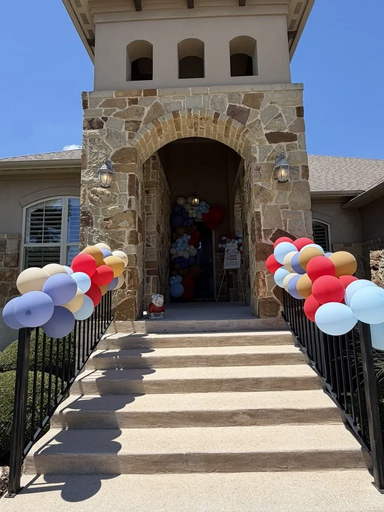 Decorated entrance with colorful balloons on black railings leading up to a stone archway on a house.
