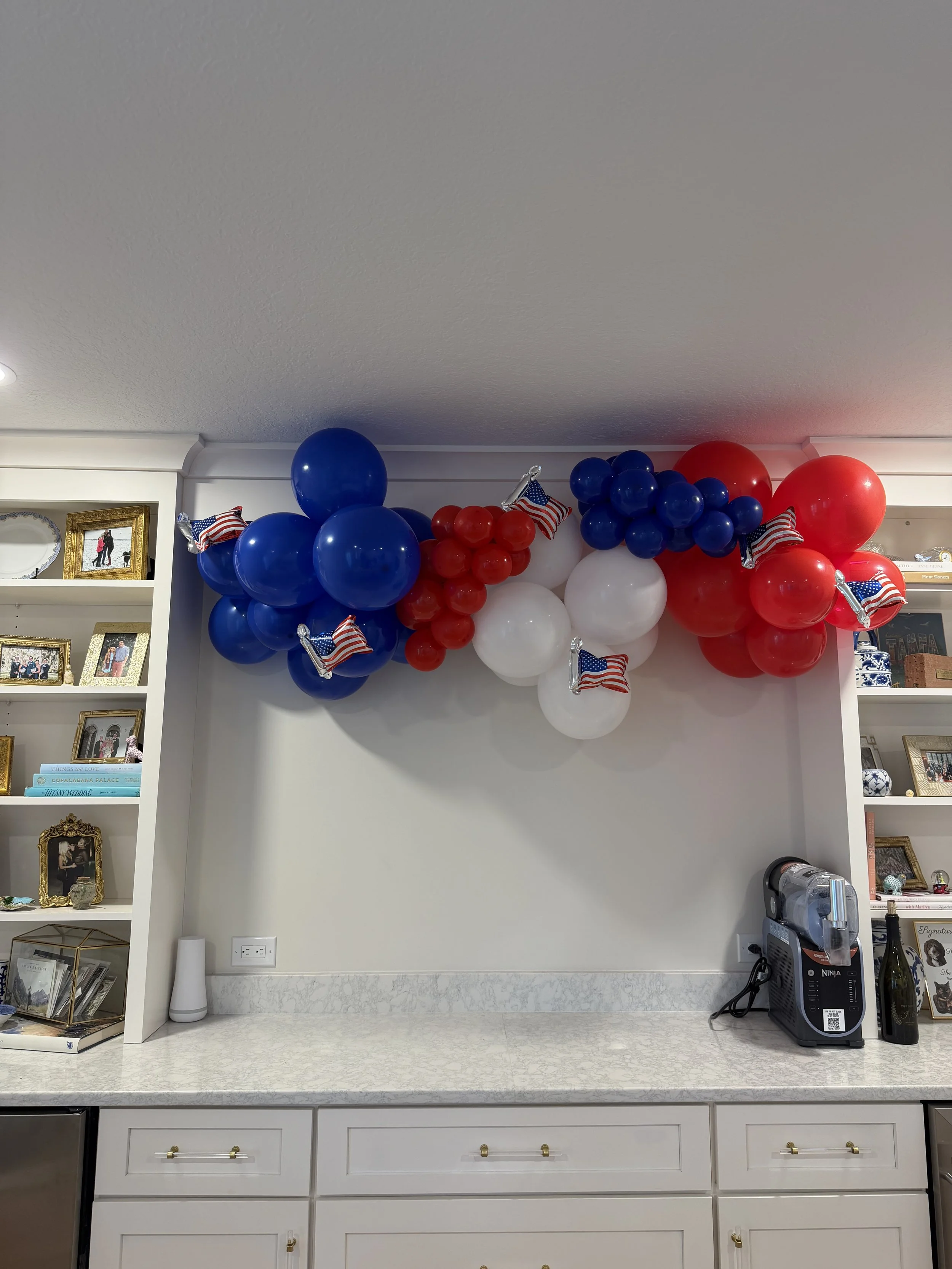 Red, white, and blue balloons with small American flags attached, arranged in a decorative arch or display on a white wall in a room, possibly for a patriotic celebration or holiday.