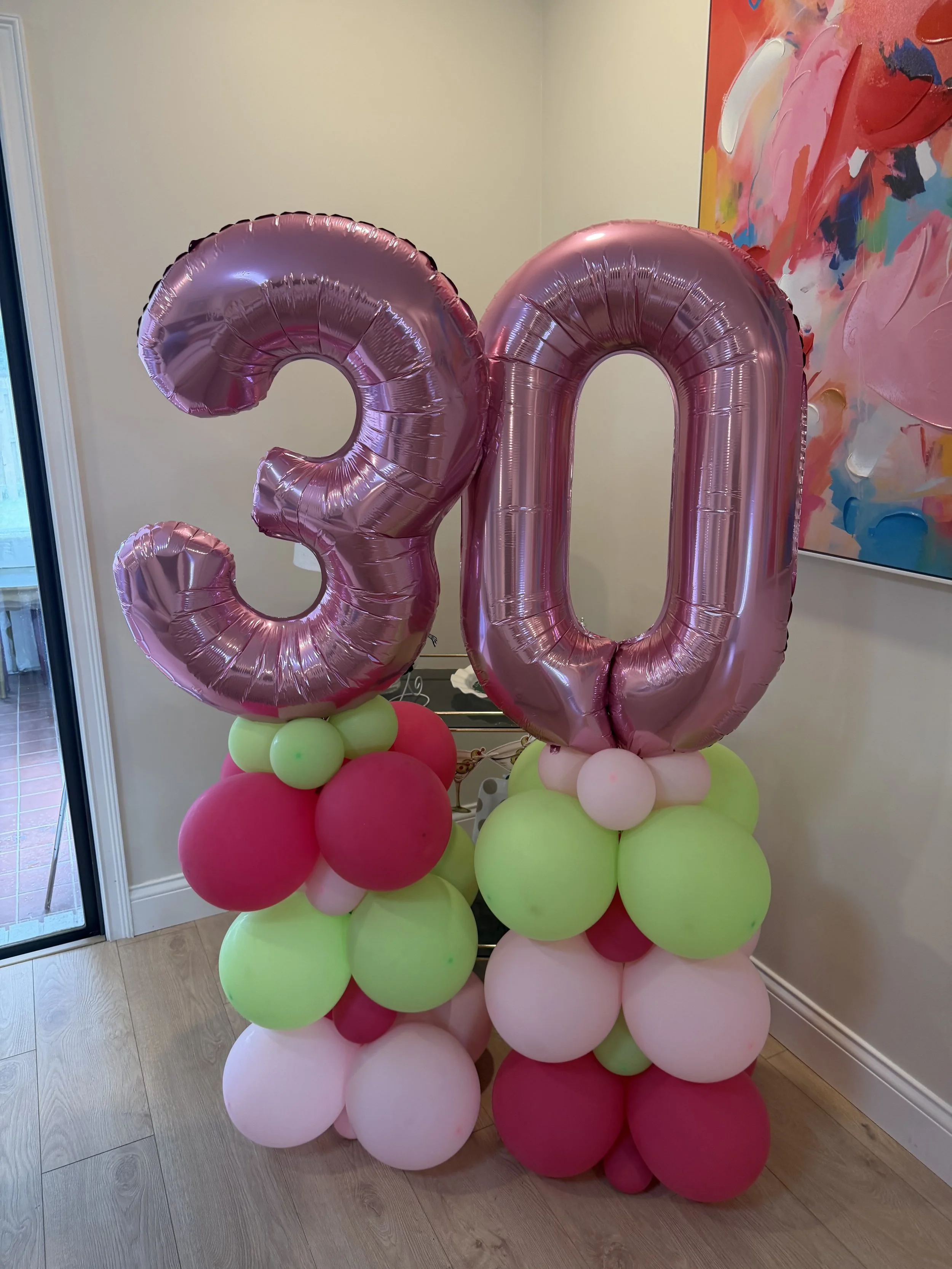 Pink number 30 balloons with clusters of pink, green, and white balloons at the base, standing near a colorful abstract painting and a window with a sliding door.