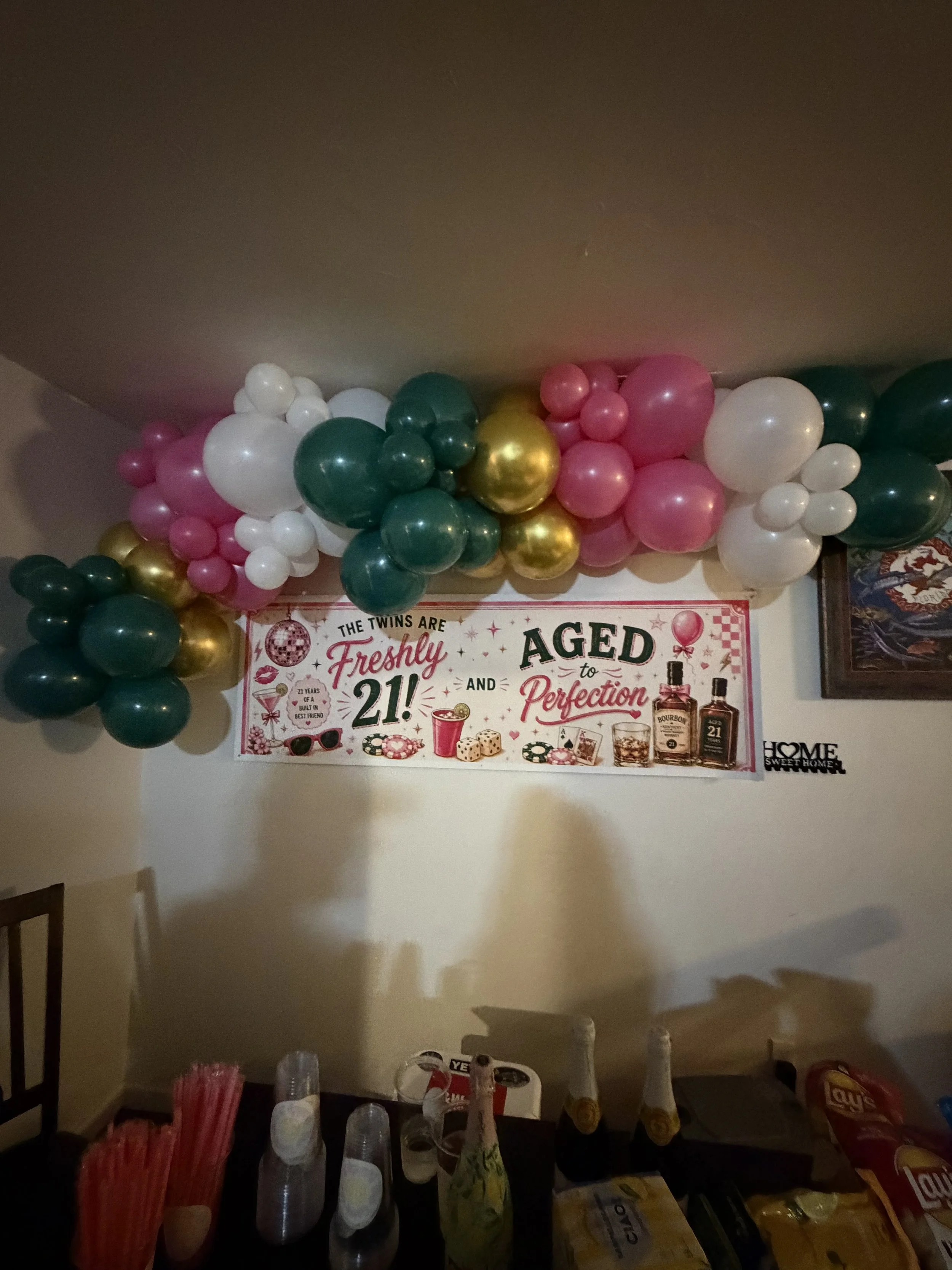Party decoration with colorful balloons in pink, green, gold, and white, arranged above a poster celebrating 21st birthdays, with party supplies on a table below.