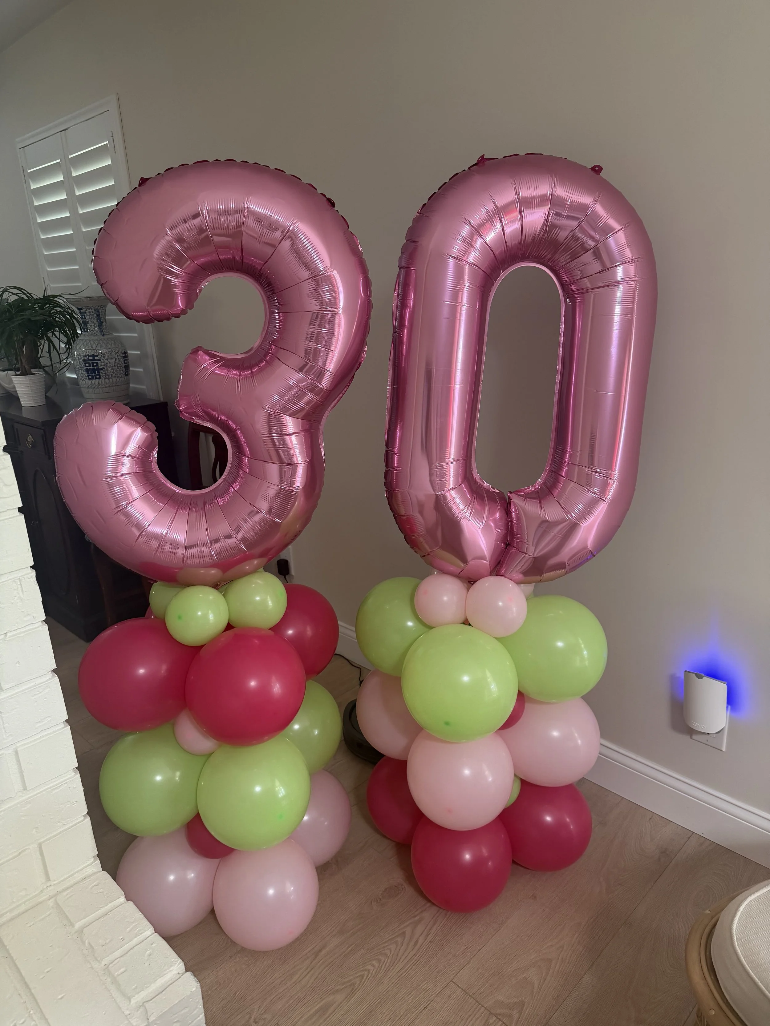 Pink number balloons '20' with colorful balloon bases, possibly for a birthday celebration, placed indoors near a white brick wall, a window with blinds, and a white speaker.