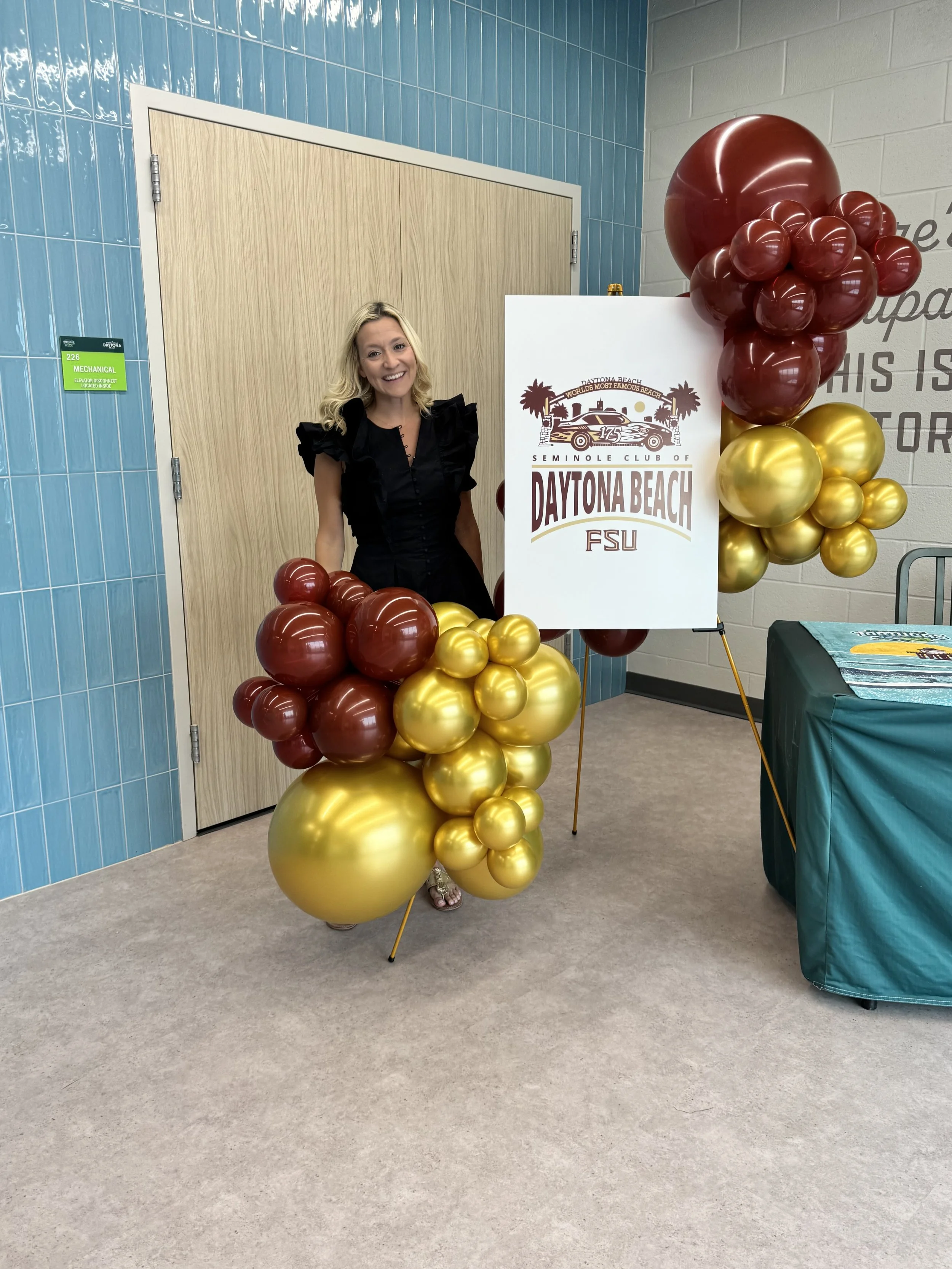 A woman in a black dress standing beside a decorative balloon display with gold and maroon balloons, and a white sign reading "Daytona Beach" and "FSU" at an indoor event.