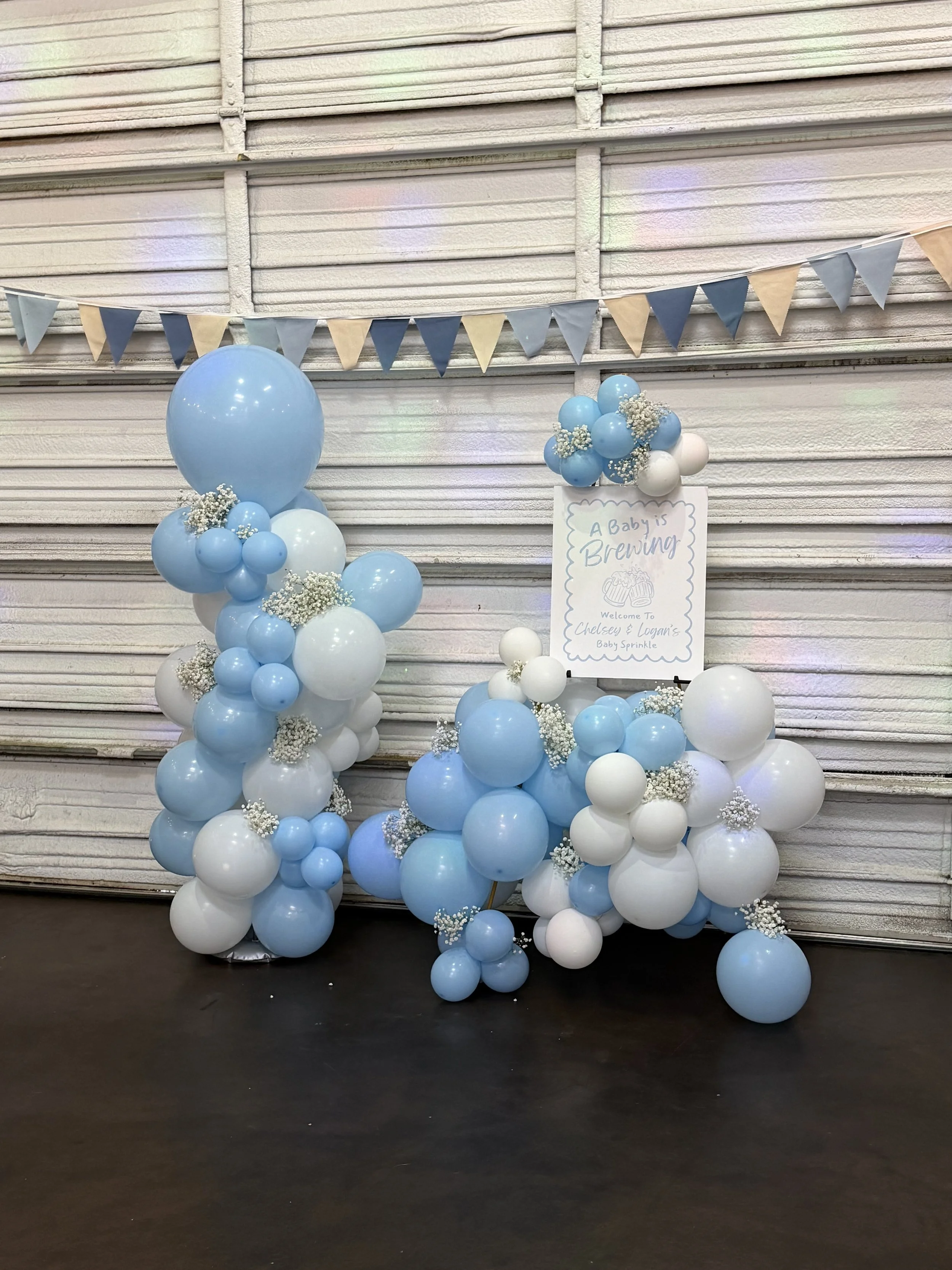 Balloon arrangement with blue, white, and light blue balloons, decorated with small white flowers, and a sign that reads "A Baby is Brewing," indicating a baby shower celebration.