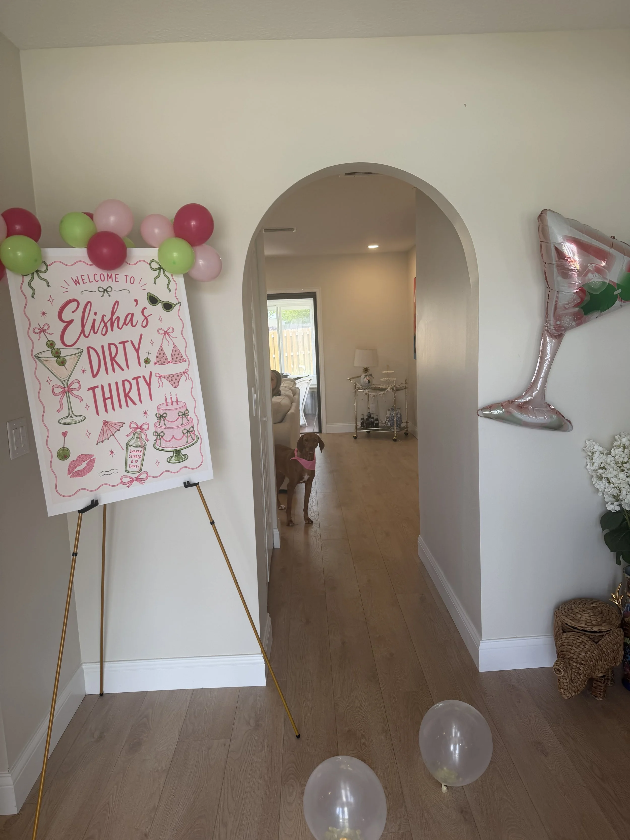 Decorative party setup with a sign that reads 'Elisha's Dirty Thirty' decorated with balloons, a large inflatable cocktail glass, and white balloons on the floor.