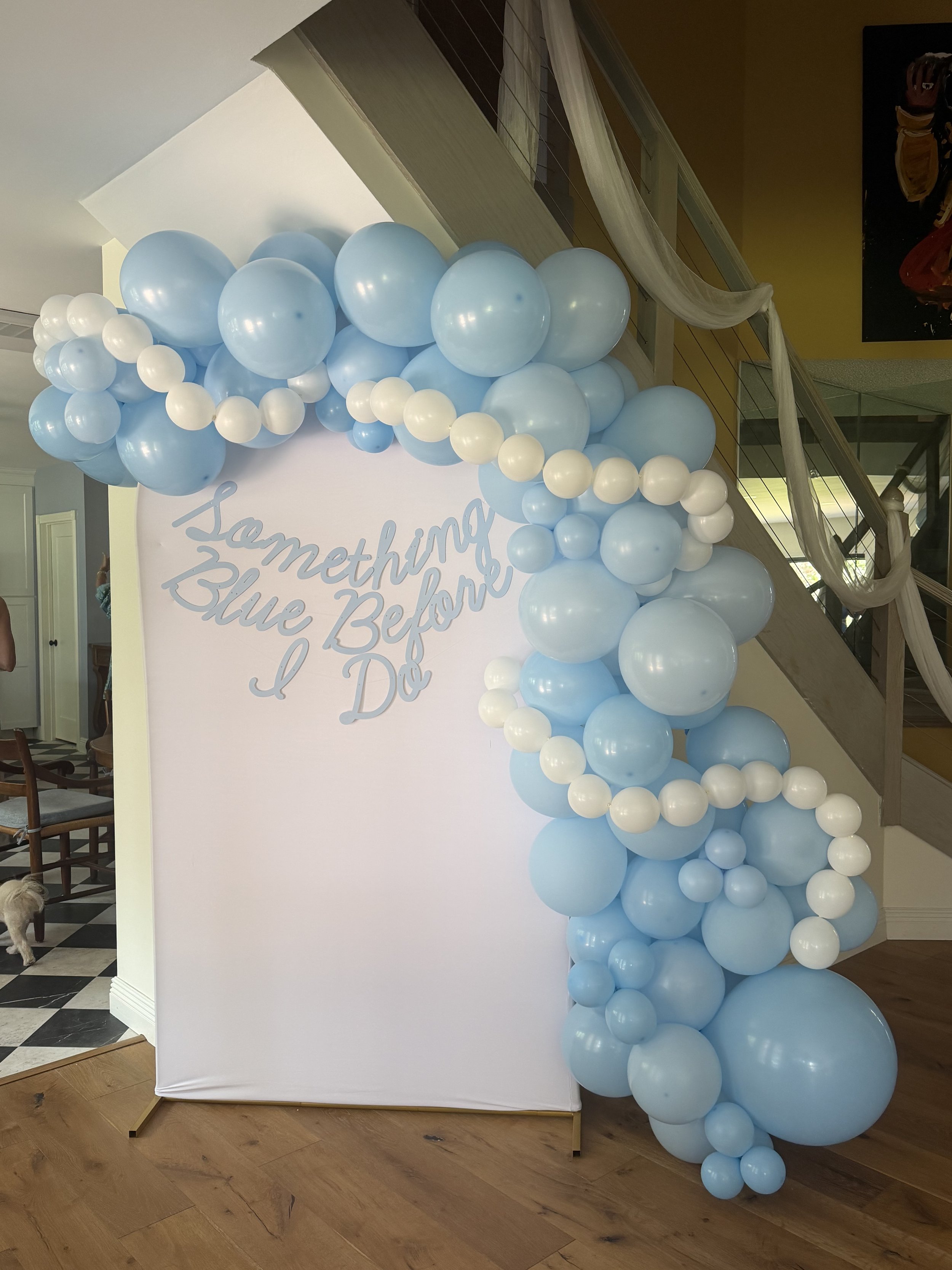 A decorative balloon arrangement with light blue and white balloons cascading from a white backdrop that has a partial phrase in cursive writing, set up indoors near a staircase.