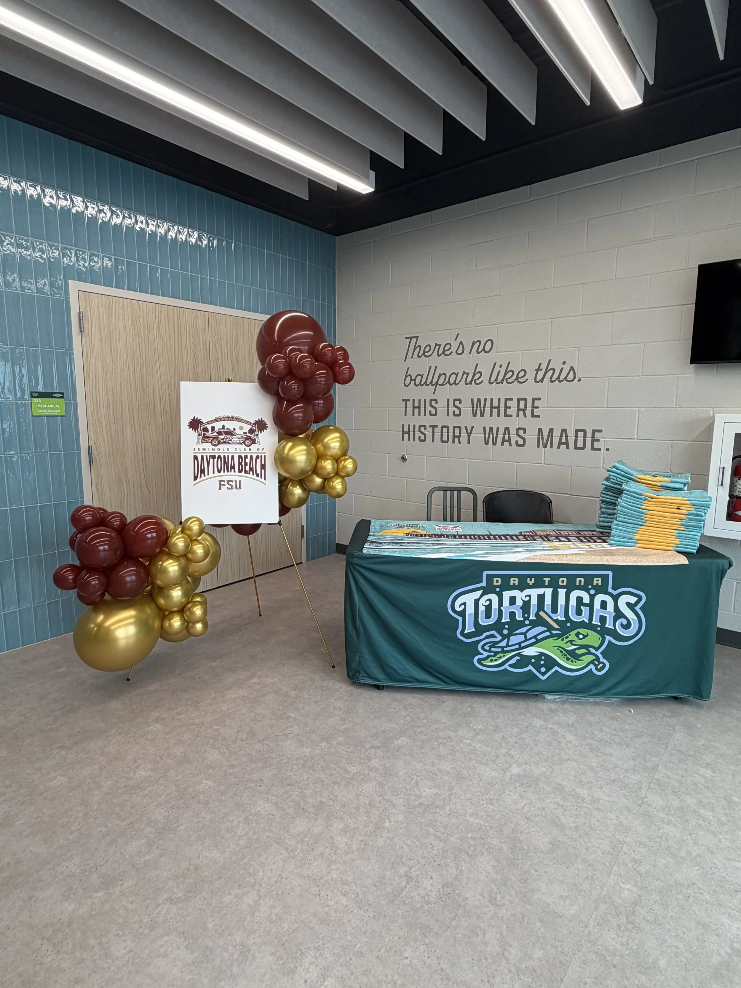 A display table with a Daytona Tortugas baseball team cover, a balloon bouquet with red and gold balloons, and a sign for Daytona Beach Florida State University. A wall features text that reads, 'There's no ballpark like this. This is where history w