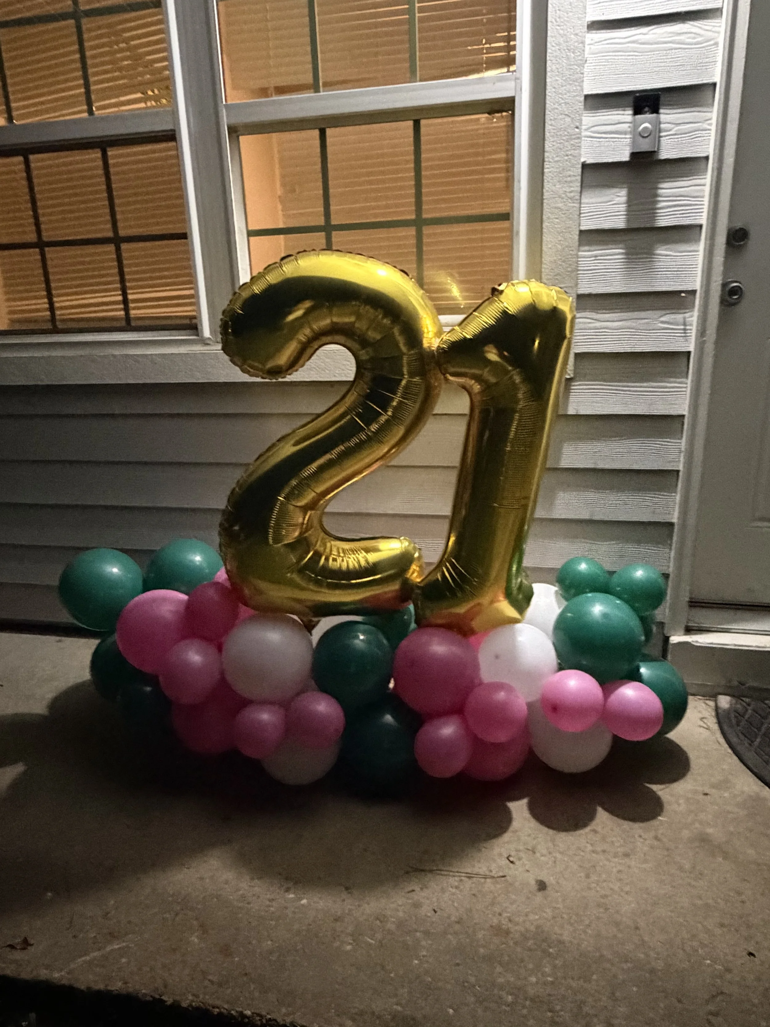 A large gold foil balloon shaped as the number 21, surrounded by pink, white, and green balloons, set against the exterior of a house with a window and siding at night.