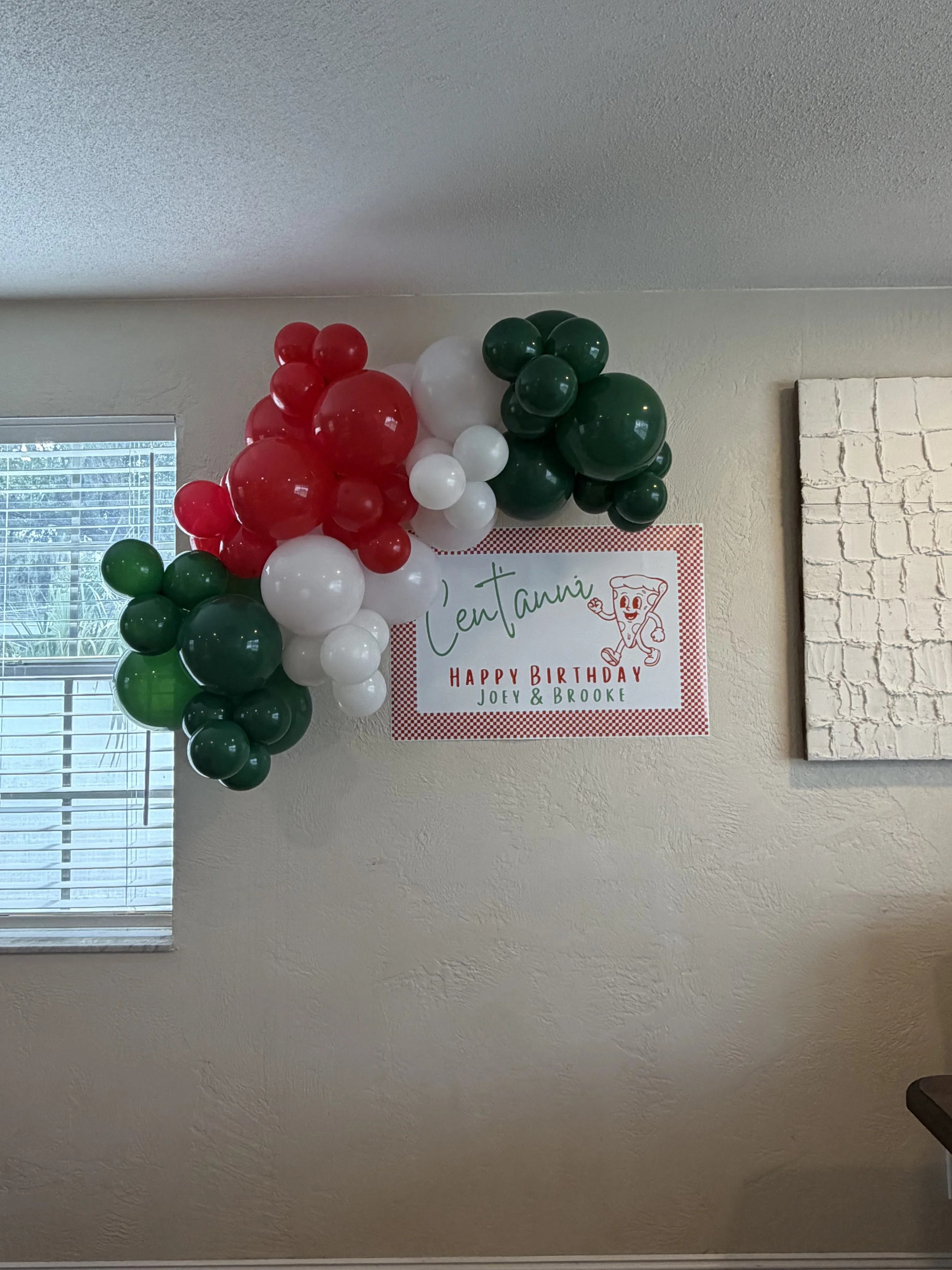 Decorative balloon arrangement in red, white, and green on a wall for a birthday celebration with a sign that reads "Tentani Happy Birthday Joey & Brooke."