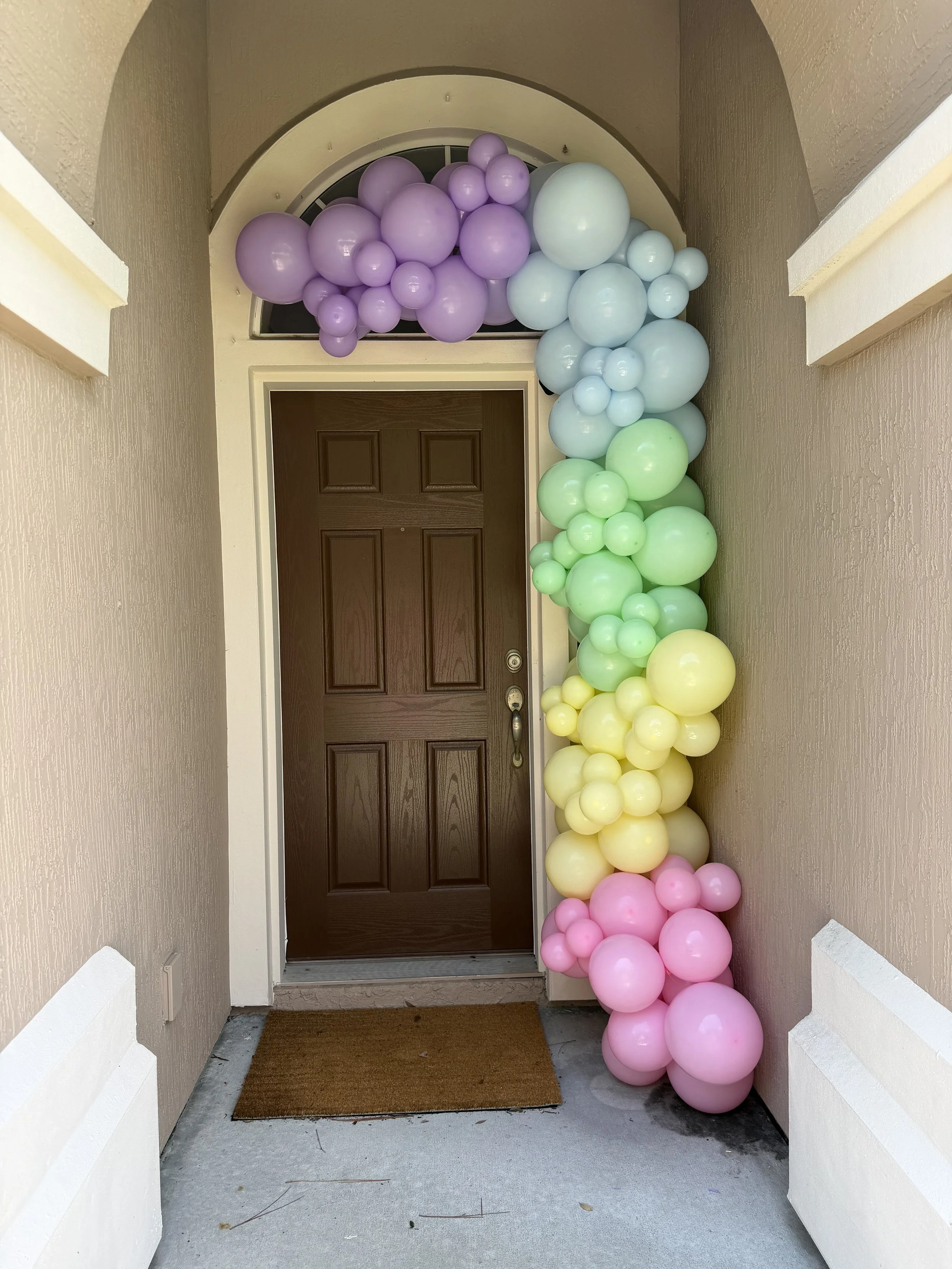Colorful balloon arrangement with pastel pink, yellow, purple, light blue, and green balloons lining a front porch and sides of a doorway.
