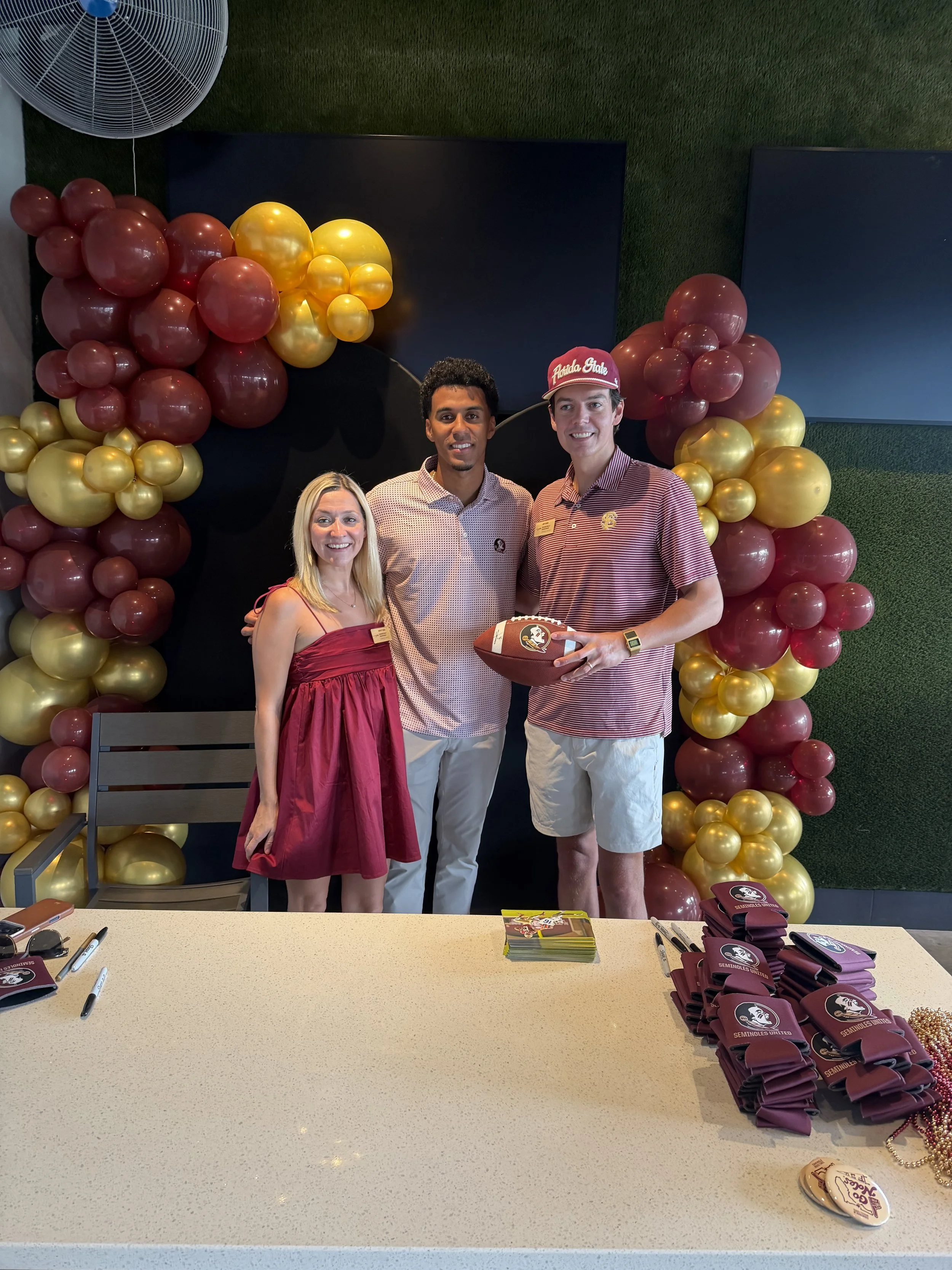 Three people standing behind a table at a university event, with balloons and university merchandise in the background.
