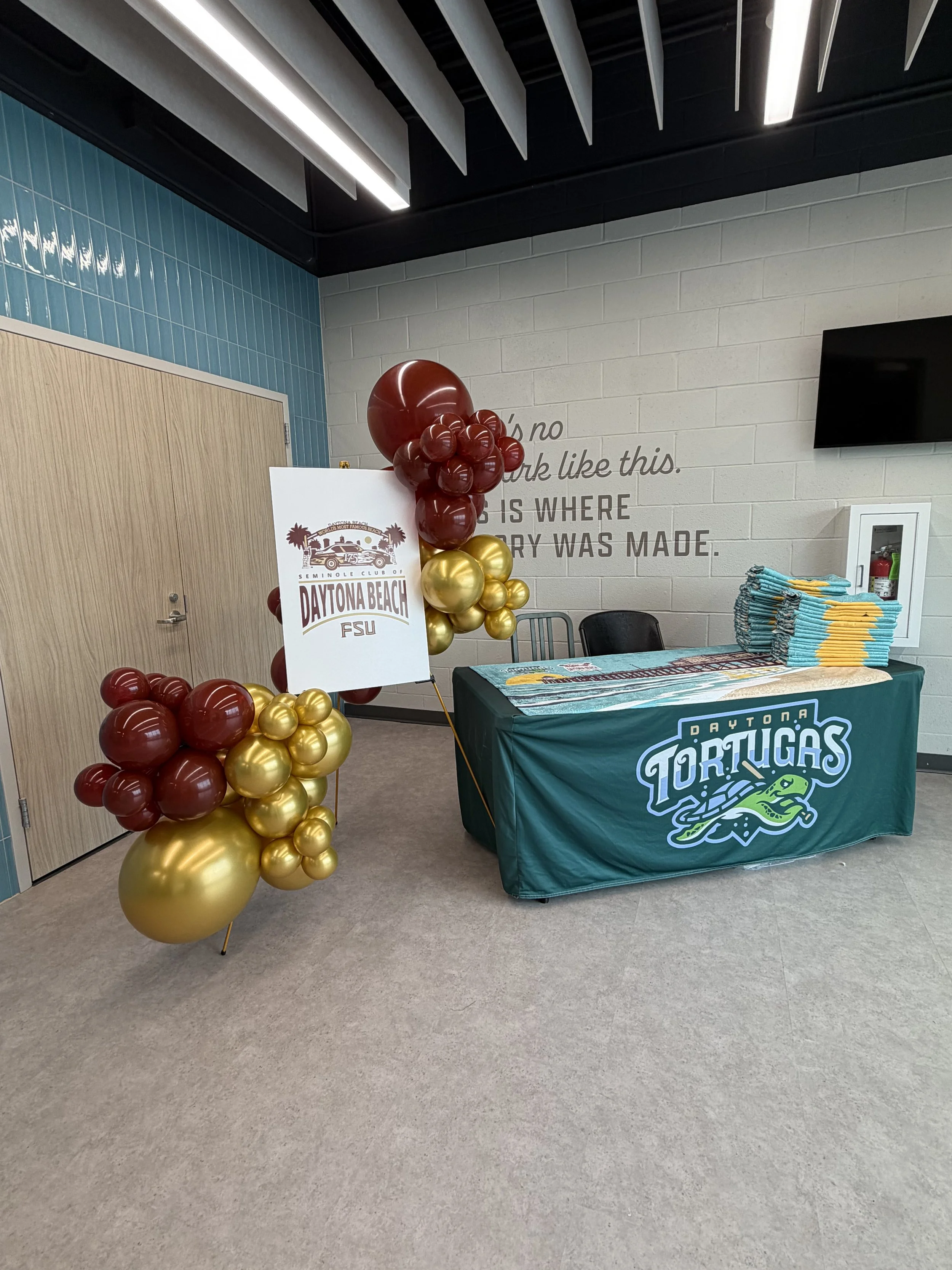 Table covered with a green tablecloth featuring the Daytona Tortugas logo, piled with stacks of folded shirts or towels, with a colorful balloon decoration and promotional sign, inside a room with white brick walls and blue tiled accent wall.