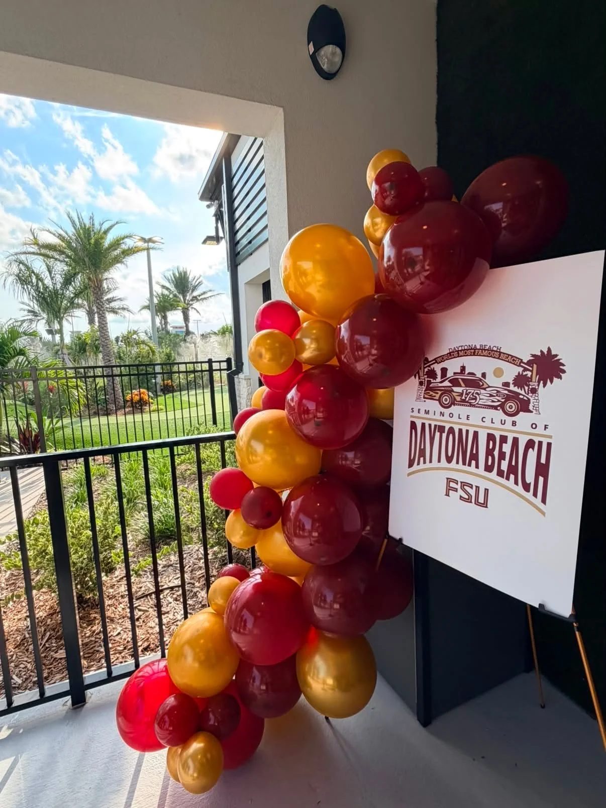 Colorful balloons in red, yellow, and gold surround a large sign for Daytona Beach Florida State University. The background shows a sunny outdoor area with palm trees and a clear sky.