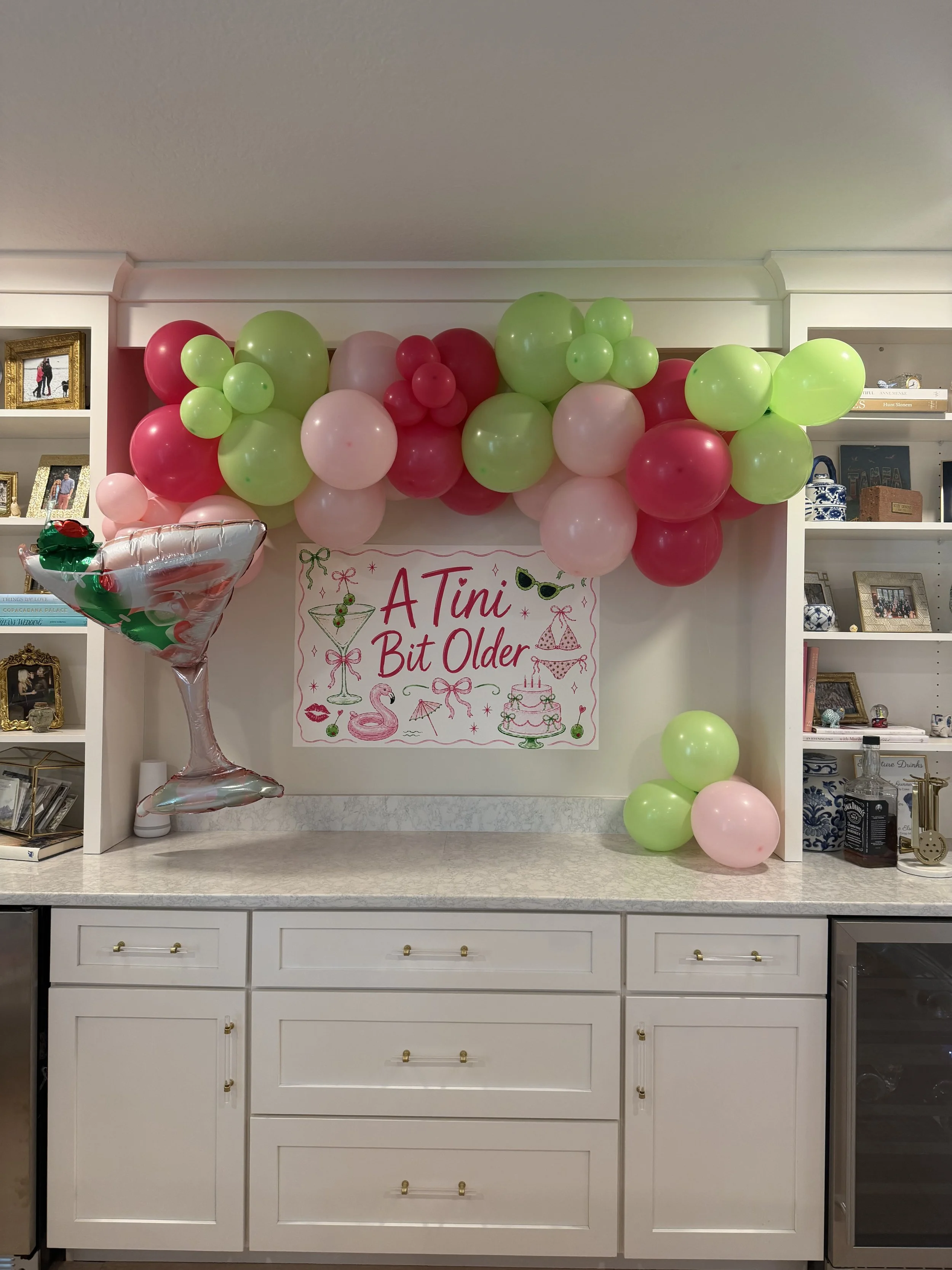 A celebration setup with pink and green balloons, a banner that reads 'A Tini Bit Older,' and a large festive cocktail balloon.