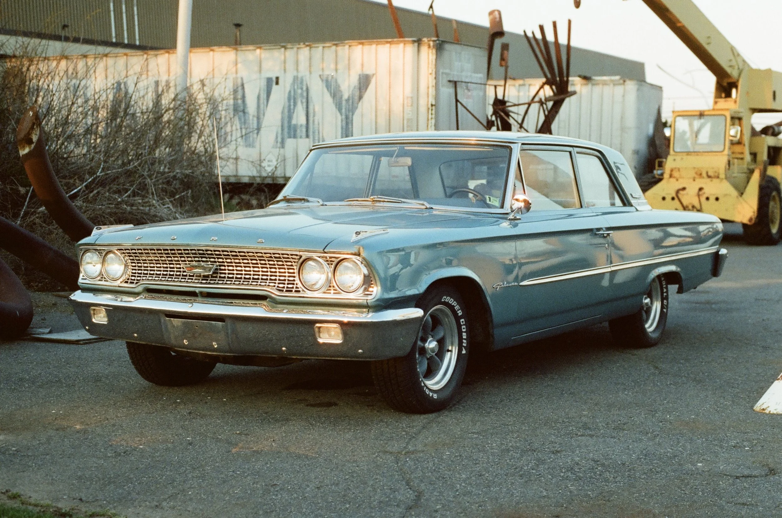 A vintage blue Ford Galaxy car parked outdoors near industrial equipment, with trees and a metal building in the background.